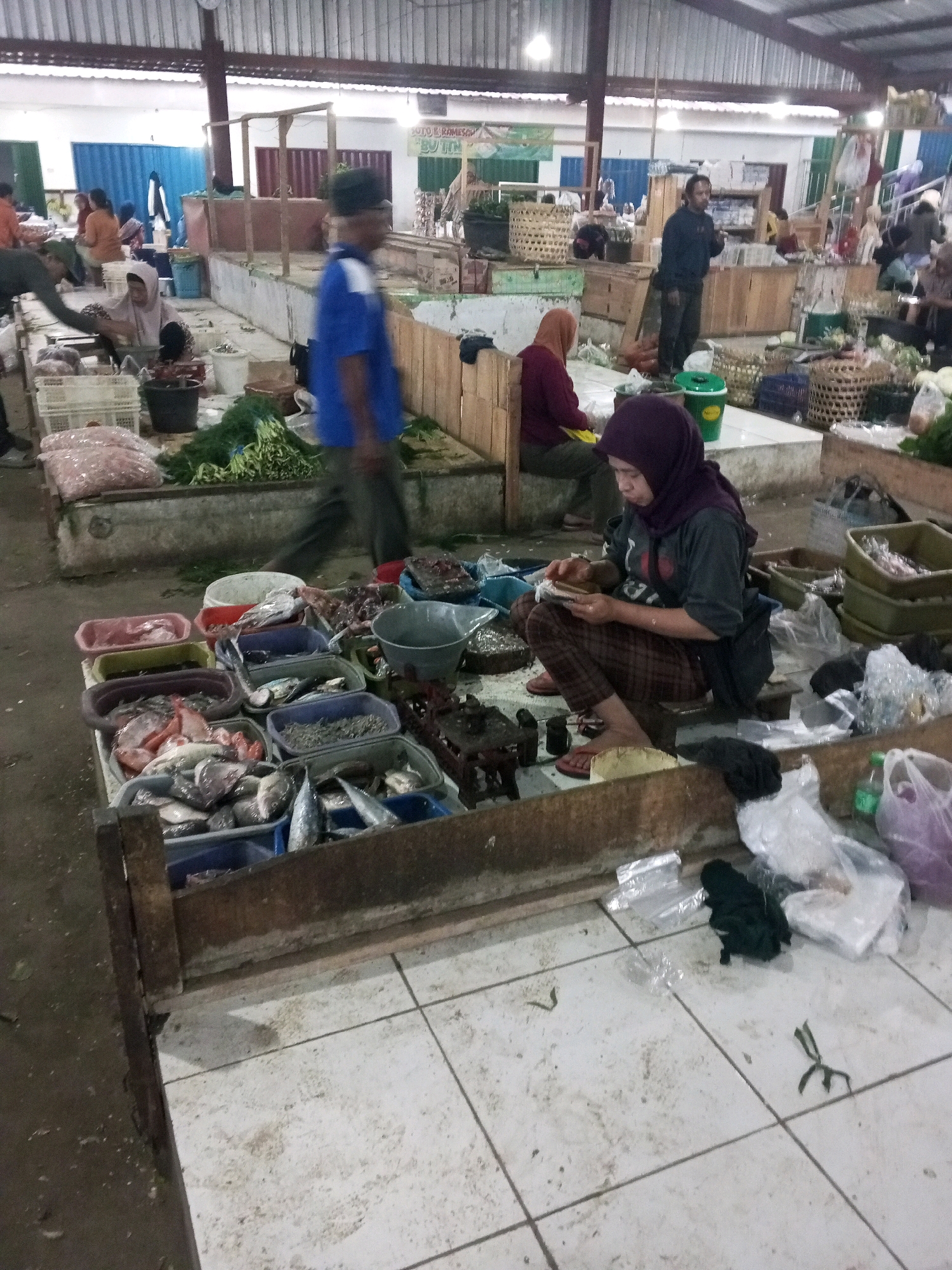 The image depicts an indoor market scene, likely in Gentan, Indonesia, with a focus on a fish stall. In the foreground, a woman wearing a purple hijab and checkered pants is seated on the floor, actively preparing fish. She is positioned in front of a wooden stall filled with various containers of fish. To the left of the stall, there are more containers with fresh produce, including what appears to be carrots and leafy greens, and bags of produce. In the background, other market-goers are visible, some tending to stalls or browsing. The environment is a covered market with a metal roof and tiled flooring. The overall impression is a bustling marketplace where fresh goods are being sold and prepared.
