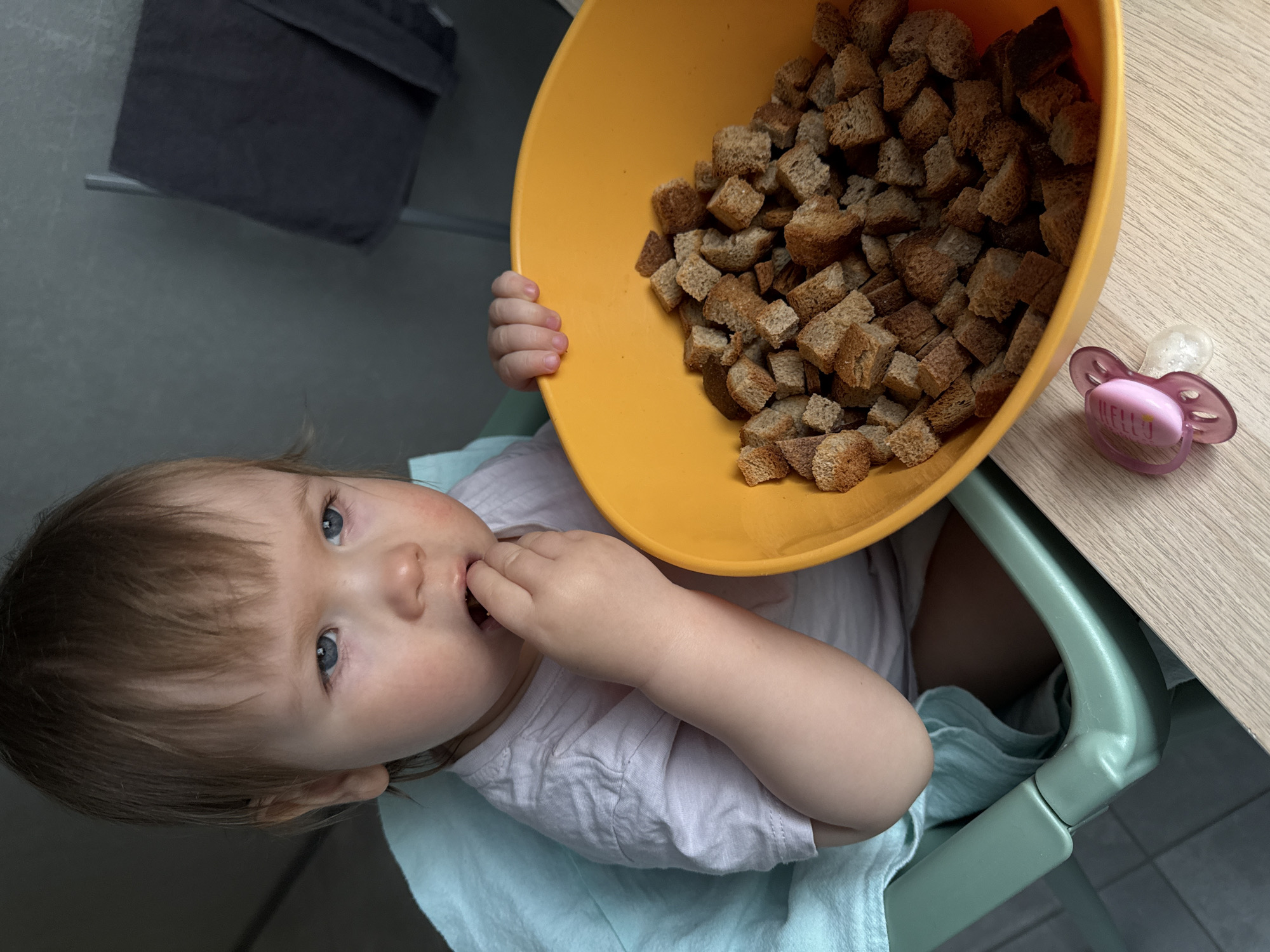 A young child, likely a toddler, is captured from a high-angle perspective, seated in a light green high chair at a light wooden table. The child has light brown hair and prominent blue eyes, looking upwards towards the camera. They are wearing a light-colored t-shirt.
One of the child's hands is near their mouth, with fingers inside, indicating they are in the process of eating or tasting something. The other hand firmly holds a large, bright orange plastic bowl that is brimming with cubed, brown bread croutons. The croutons appear well-toasted and are the central focus of the bowl.
On the wooden table, to the right of the bowl, rests a pink pacifier with a clear silicone nipple. The word "HELLO" is clearly visible in white lettering on the pink part of the pacifier.
The background is simple, featuring a muted grey wall or floor in the lower left and a dark grey towel hanging on a rail in the upper left, suggesting an indoor domestic setting. The photograph captures a candid moment of the child during snack time in Moscow, Russia.