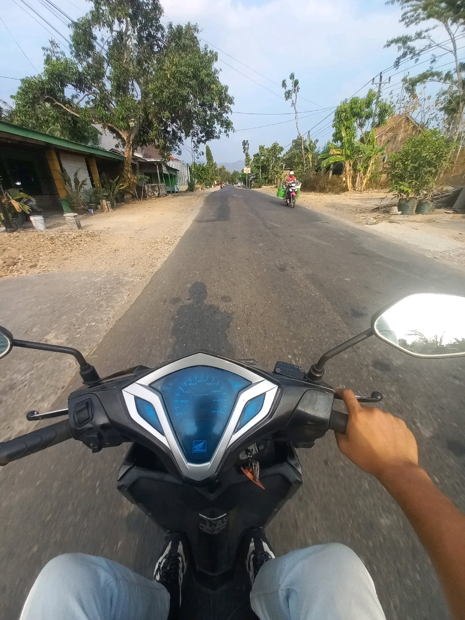 The image depicts a first-person perspective of riding a motorcycle on a road in Ngarjosari, Indonesia. The road is paved, with a gravel shoulder on the left side. On the left side of the road, there are buildings and trees, and on the right side, there are more trees and some structures. The sky is clear and blue. In the foreground, the motorcycle's dashboard, handlebars, and a hand are visible. The dashboard displays speed in kilometers per hour, with a blue digital display. Further down the road, another motorcycle is visible with a rider wearing a red shirt and a white helmet, carrying something on the back. The overall scene is rural and sunny.