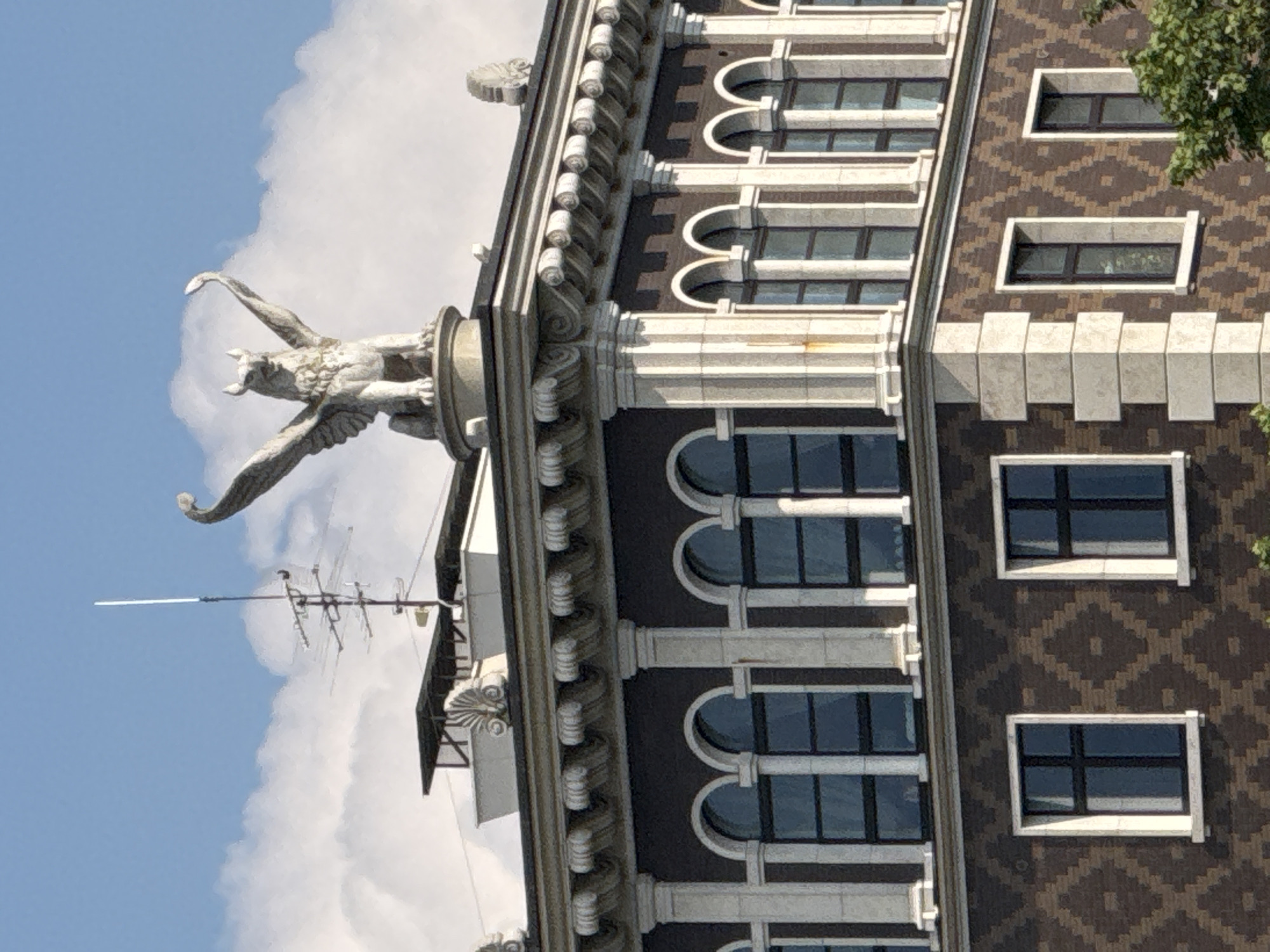 The image shows the facade of a building with ornate architectural details. A statue of a winged creature, possibly a griffin, is visible on the left side of the building, silhouetted against a bright blue sky filled with white clouds. The building itself is a multi-story structure with a dark brown tiled exterior featuring a diamond pattern. Rows of windows with white frames are visible, some rectangular and some arched. Decorative elements like columns and moldings adorn the facade. On the left side of the image, an antenna is mounted on the building. The image appears to be taken during daylight, with clear weather conditions. The overall impression is one of grandeur and historical architecture.