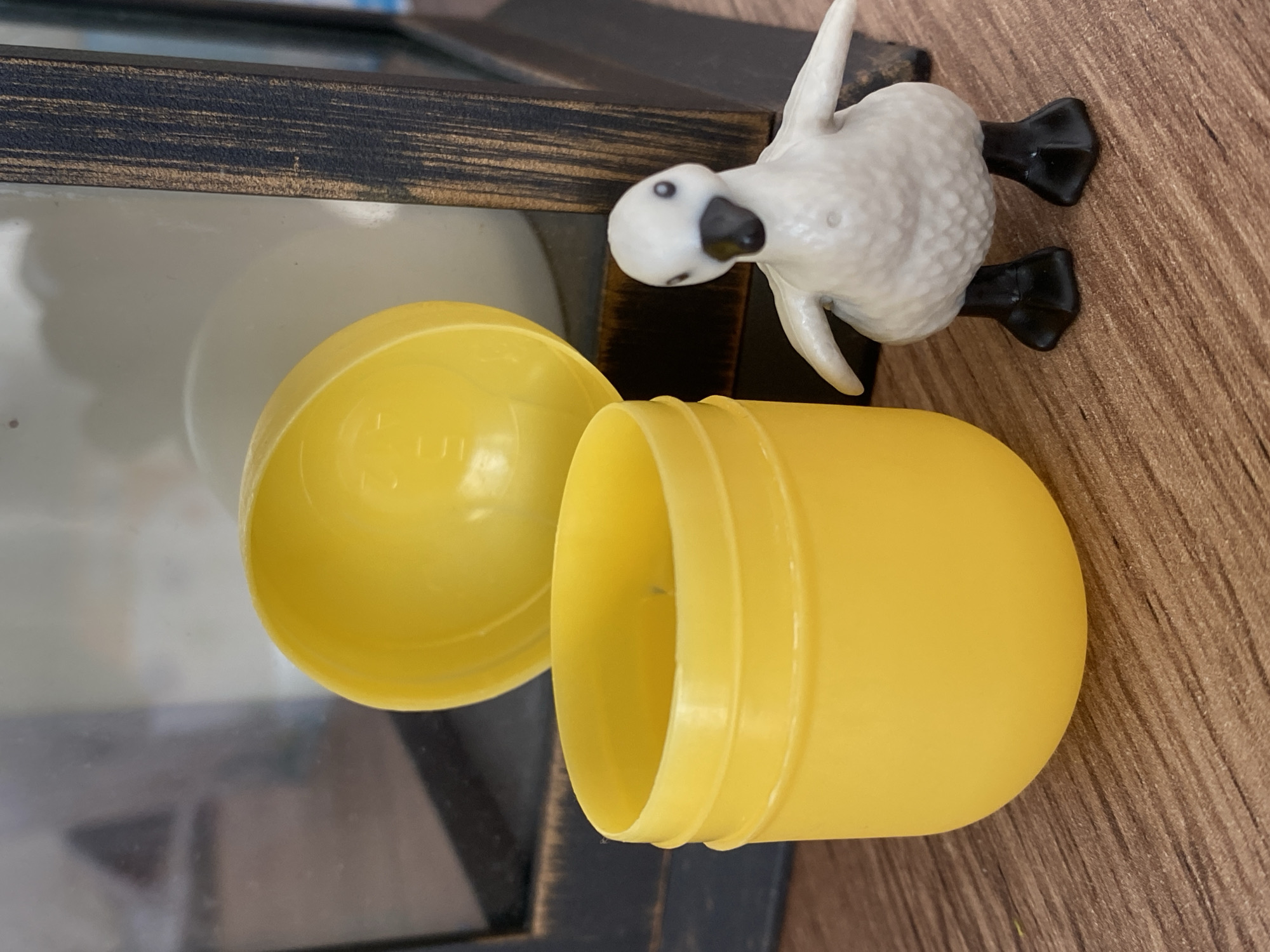 A white toy duck with black feet stands on a wooden surface next to an open, yellow plastic egg. The duck is facing left, with its head tilted slightly down. Its wings are raised, and its body is textured to resemble feathers. The egg is split into two halves, with one half, the lid, lying to the left of the other half, which is upright. The egg has a smooth, rounded shape and a ribbed rim where it would close. Behind the duck and egg is a dark, rectangular frame with a distressed wood grain texture. Within the frame, a blurry, light-colored object is visible, possibly a mirror or glass surface reflecting light. The scene is lit from above, casting soft shadows. The overall impression is one of a child's toy on a table or shelf. There is no visible text in the image. The time of day, weather, and specific location are not discernible.