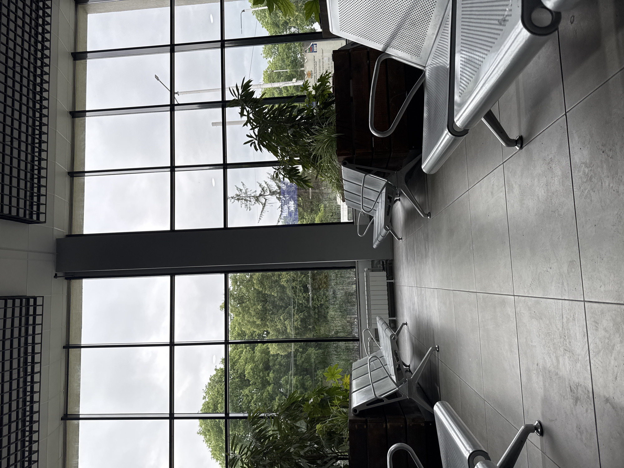 The image captures an interior space, possibly a waiting area or transportation hub, in Pleven, Bulgaria. The scene is characterized by large windows offering a view of the outside, revealing overcast skies and lush greenery including trees and bushes. The window structure has dark framing, creating a grid pattern.
Inside, rows of metal seating with perforated surfaces are arranged. These seats are mounted on the floor and are in rows. Plants in dark wooden containers are nestled behind the seating. The floor is covered with large square tiles in gray.
The exterior visible through the windows includes a glimpse of buildings and street infrastructure such as lampposts. There is little indication of human presence in the image, focusing instead on the architectural and design elements of the space. A framed grating design is on the left wall.