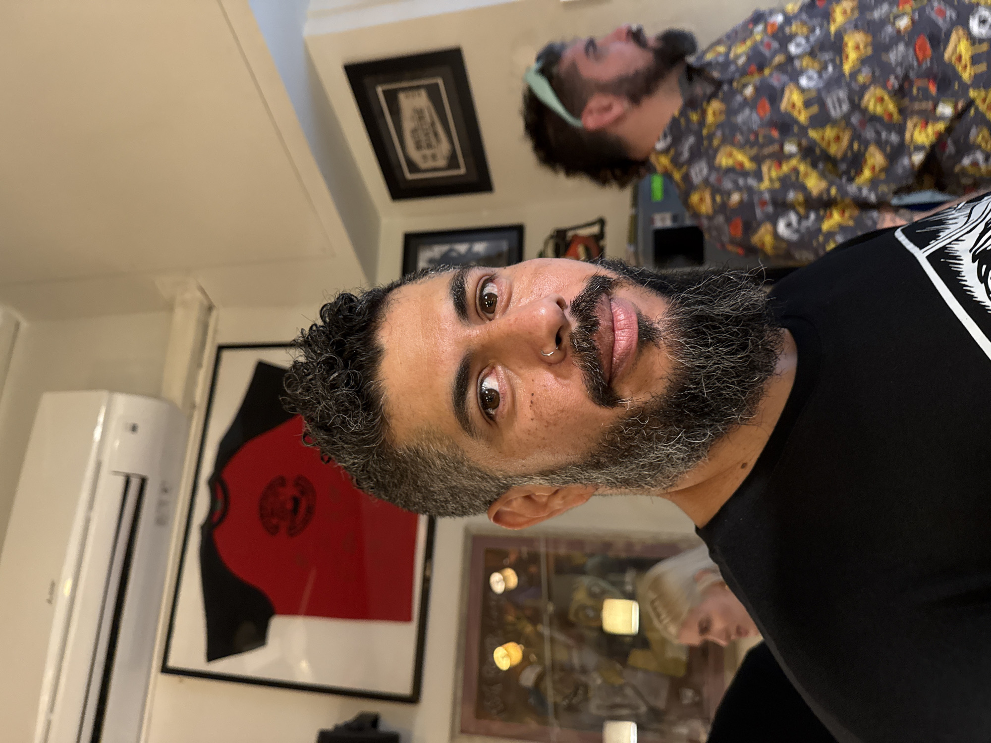 A close-up indoor shot depicts a man with salt-and-pepper curly hair, a dark beard, and a nose ring, wearing a black t-shirt with a white print. Behind him, another man is wearing a patterned shirt with construction vehicles and food. Framed art adorns the walls, including a red and black shirt in a frame and other decorative prints. An air conditioning unit is visible to the left. The location is likely indoors but not identifiable as Barcelona or any specific city.