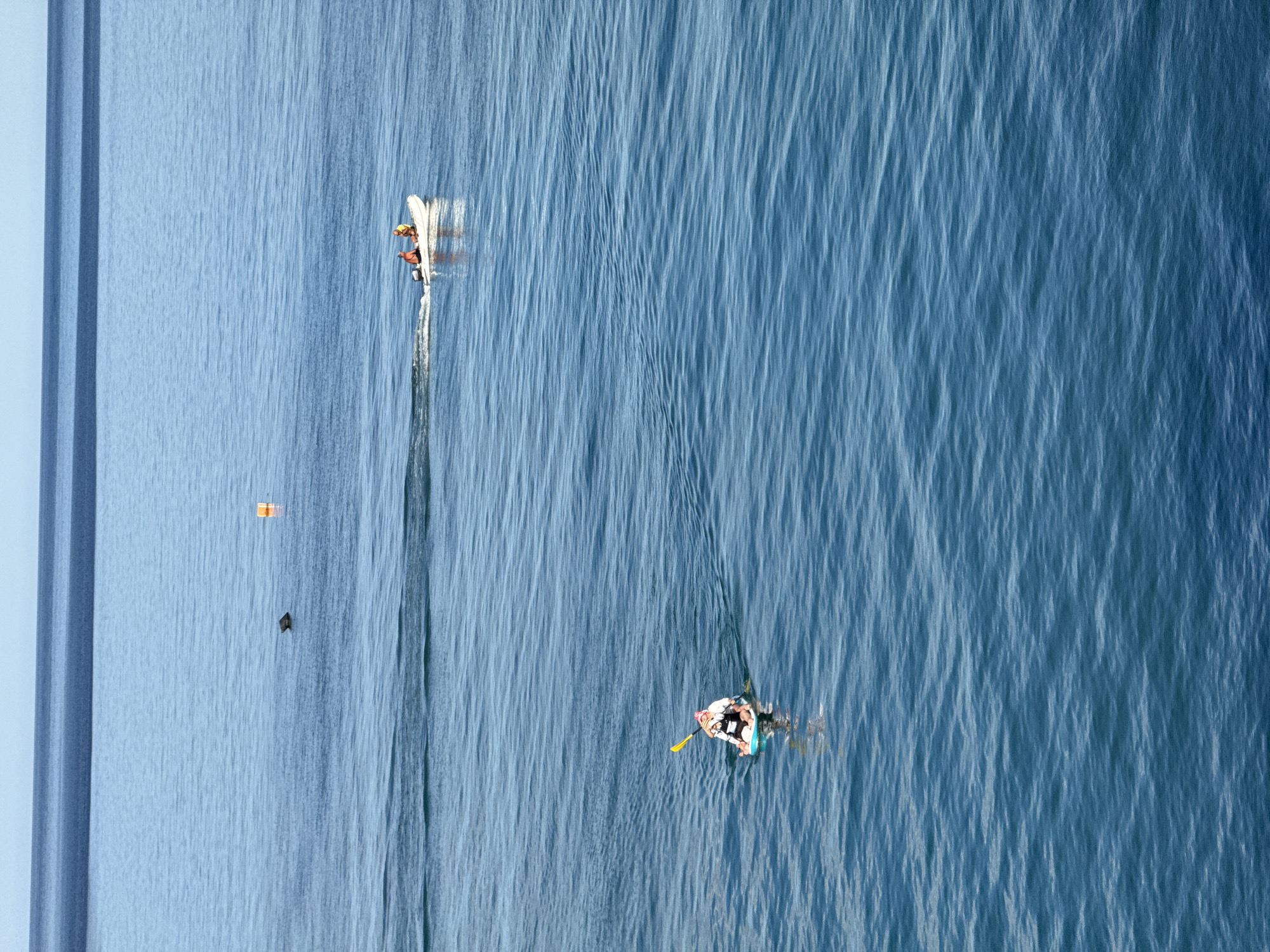 The image shows a wide expanse of blue ocean with gentle waves under a clear sky. In the distance, two people are in a white inflatable boat with an outboard motor, moving from left to right. Further left and closer to the foreground, a person is sitting on a paddleboard, holding a yellow paddle. There are also a few buoys visible in the water. The scene depicts a calm day at sea.