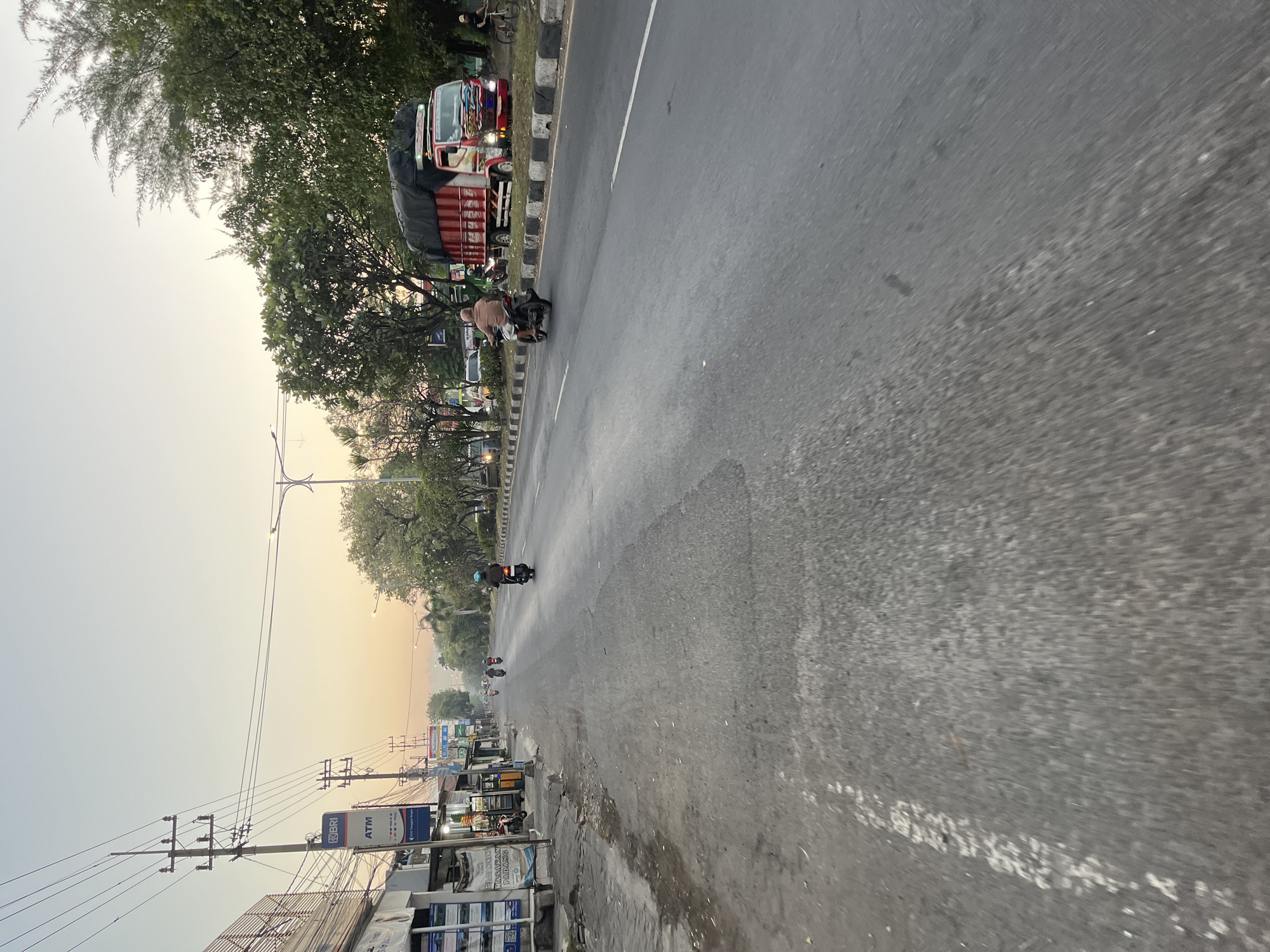 This is a street-level photograph taken in the city of Ngaban, Indonesia, likely during the early morning or late afternoon, given the soft, warm light. The image depicts a two-lane asphalt road with traffic. On the left side of the road, there are several shops and businesses, including one with a prominent "BRI ATM" sign. Utility poles with many wires are visible along this side of the street.
Several motorcycles are present on the road. One motorcycle with a rider wearing a brown shirt is in the foreground, moving away from the camera. Further down the road, other motorcycles are also in motion. On the right side of the road, a red and white truck with a tarp-covered load is stopped or moving slowly. A person on a bicycle is also visible near the truck. Trees line the right side of the road, with some foliage visible in the background. The sky is clear and has a gradient of light colors, suggesting sunrise or sunset. The overall atmosphere is that of a typical urban street scene in Indonesia.