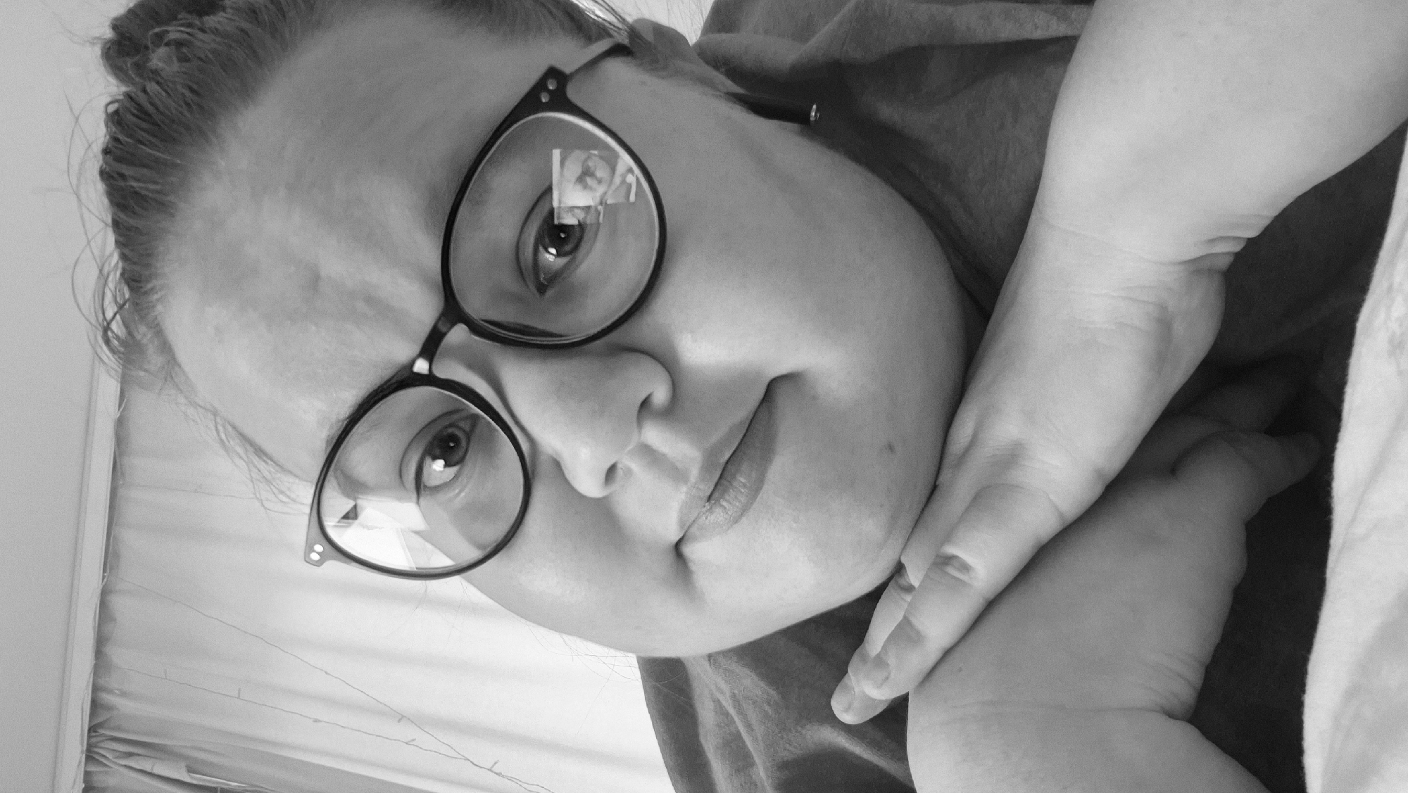 A black and white, close-up selfie depicts a woman wearing glasses with a dark, thick frame. Her face is positioned at a slight angle, with her right hand gently supporting her chin. Her hair is pulled back in a casual style. The setting suggests an indoor environment, possibly a domestic space, as indicated by the bedding visible in the lower right corner of the frame. There is no information or indication that this image was taken in the city of Chaikovsky, Russia.