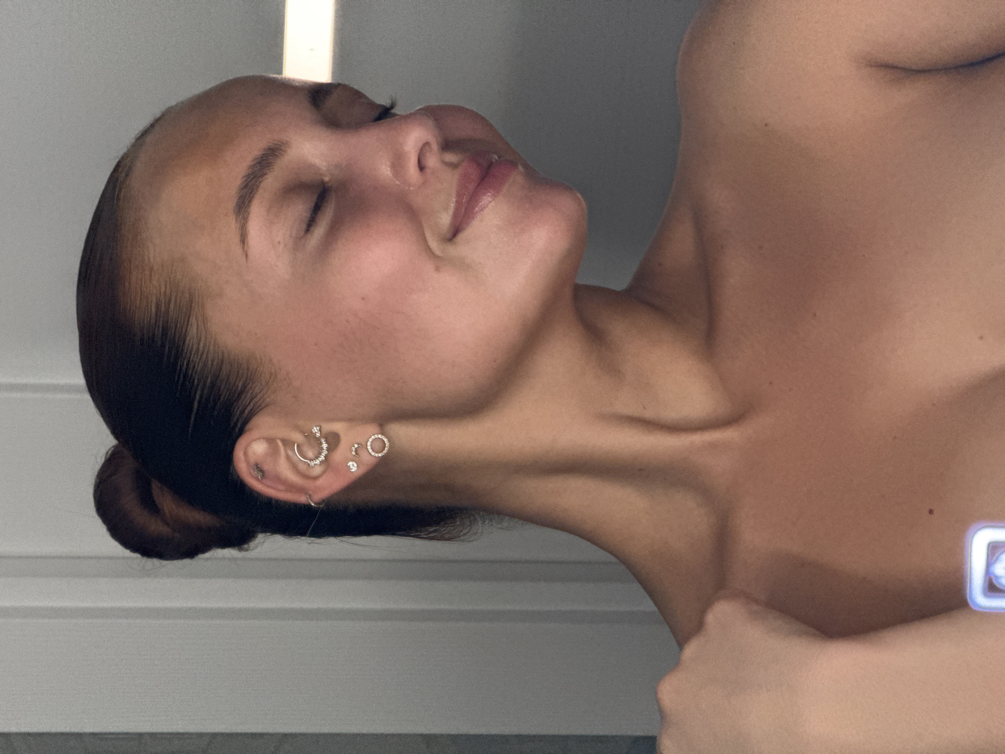 The media file presents a close-up portrait of a person, likely female, captured from the chest upwards. The image is oriented sideways, suggesting the subject is lying down or tilting their head significantly. The person has their eyes closed and a soft, serene smile, conveying a sense of peace and relaxation. Their dark hair is neatly pulled back into a bun at the nape of their neck.
The subject's right ear is adorned with multiple piercings, including a prominent hoop earring encrusted with small stones, several stud earrings, and another small hoop. Their skin appears to have a warm, tanned complexion, with a few small moles visible on the neck and shoulder. The lighting is soft but bright, highlighting the contours of their face and collarbone.
The background is simple and uncluttered, consisting of a light-colored, possibly paneled, wall or vertical surface. A bright vertical strip of light is visible in the upper background, suggesting a light source or a window. A hand is partially visible in the lower right corner, resting on the subject's shoulder/chest area, possibly indicating a selfie or a close-up shot. There is no visible text in the image. While the location context is Chaikovsky, Russia, there are no specific visual cues within the image to confirm this. The overall mood is one of tranquility and quiet contentment.