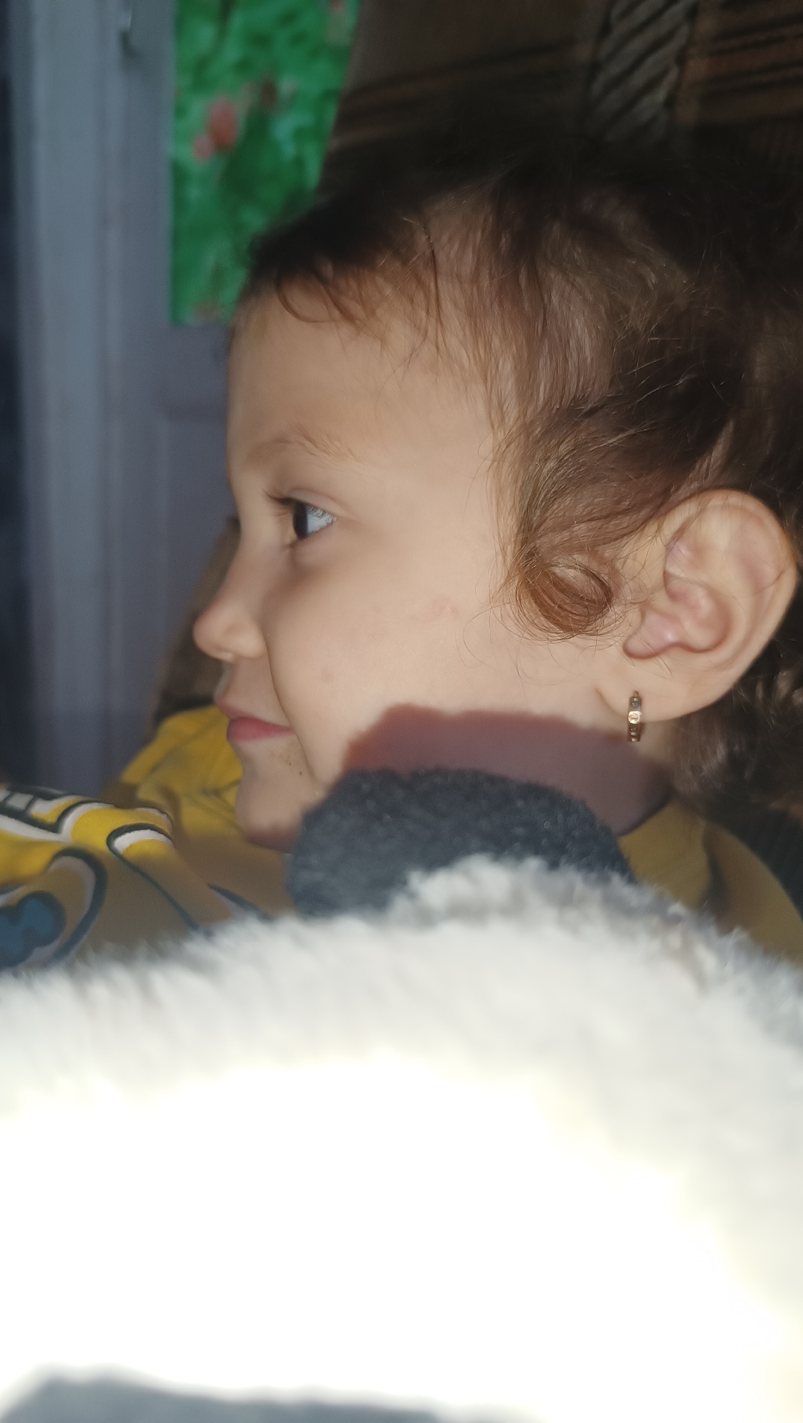 A close-up, side-profile shot shows a young child with light brown hair and fair skin. The child is wearing a yellow shirt with blue accents and a small gold hoop earring in their left ear. The child is looking to the left of the frame. In the foreground, a blurred, fluffy white object partially obstructs the lower portion of the image, and a dark gray fuzzy object is visible behind it. The background is out of focus and shows a hint of a green and red image on the wall and a textured surface above it. There is no indication of the city of Karsha, Russia, or any urban environment in this image.