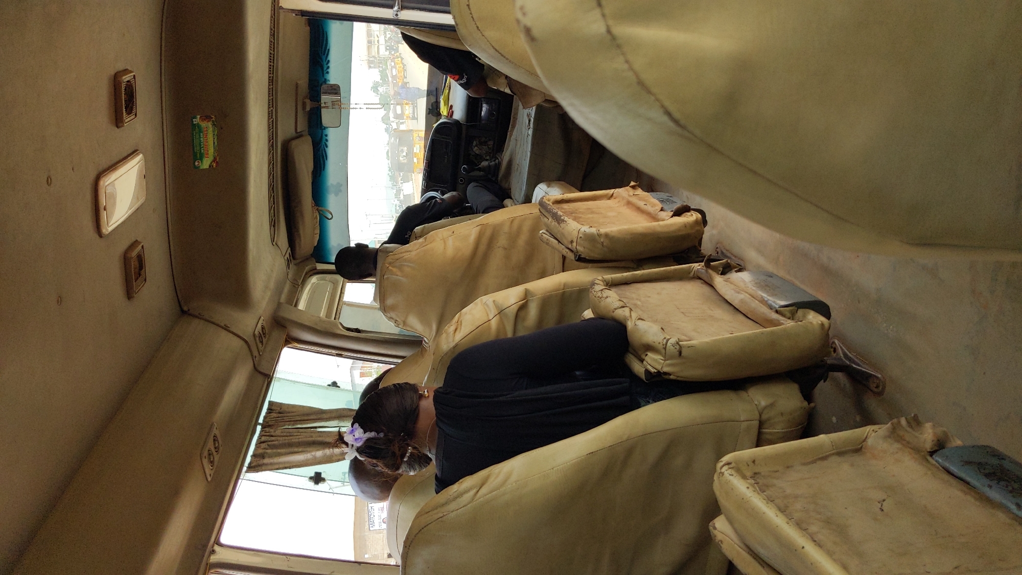 This image, presented on its side, provides a view inside a public transport vehicle, likely a bus or large minivan, looking towards the front. The interior features rows of utilitarian seats upholstered in a worn, light beige or tan material, with some showing exposed frames and padding.
Several passengers are visible, seated and facing forward. In the foreground, a person wearing a black top is seen from behind, their hair tied back with a light-colored scrunchie. Further down the aisle, another individual in a dark shirt is seated. A driver or front passenger is partially visible near the windshield.
The vehicle's ceiling is light-colored and fitted with rectangular light fixtures or vents. A small, colorful packet, with "Bournvita" partially legible, is attached to the ceiling. Through the side windows, a bright daytime outdoor scene is visible, showing glimpses of other vehicles and an urban or semi-urban environment. Light-colored curtains are drawn on some of the windows. A sign or sticker seen through a window on the lower right corner displays "MATOKH" and "PRICES," offering a local detail.
The scene depicts routine travel within a functional, well-used public conveyance. No specific activities or emotions are clearly discernible from the passengers, who appear to be simply commuting.