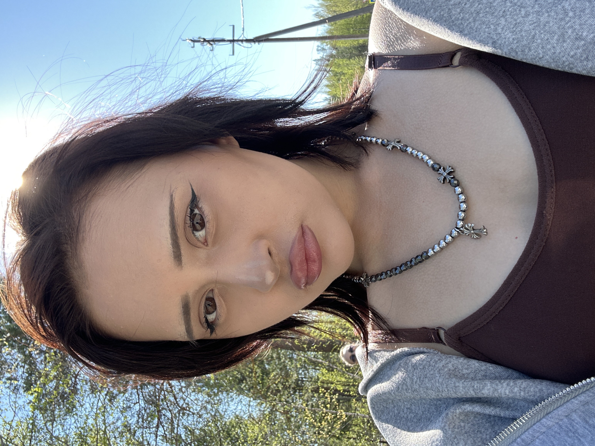A young woman with dark hair and makeup, wearing a brown top and gray zip-up hoodie, is positioned outdoors in a natural environment under a bright sky. She wears a cross necklace. Overhead power lines and trees suggest a location possibly on the outskirts of a city. The image captures a selfie-style close-up. This location is not recognizable as Высокий, Russia from the provided image.