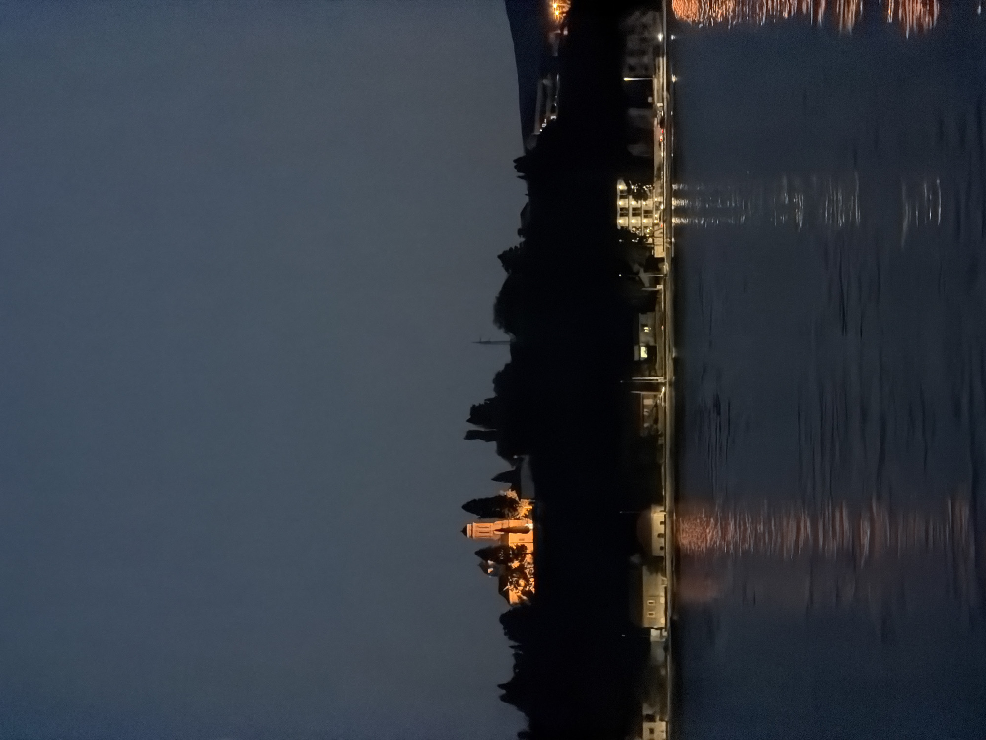 The image depicts a nighttime scene of the Bay of Kotor near Đurići, Montenegro. The foreground is dominated by a dark body of water, likely the bay, with rippling reflections of lights from the shore. In the middle ground, a silhouetted hillside rises, upon which several structures are visible. A prominent building, illuminated with warm, orange light, stands out on the crest of the hill, featuring a distinct tower and dome. This building appears to be a church or monastery. Scattered trees are also silhouetted against the dark sky. Along the base of the hillside, near the water's edge, are several smaller, illuminated buildings, suggesting a coastal village or settlement. The sky above is a deep, dark blue, typical of nighttime. The overall impression is one of serene, illuminated architecture against a dark natural landscape by the water.