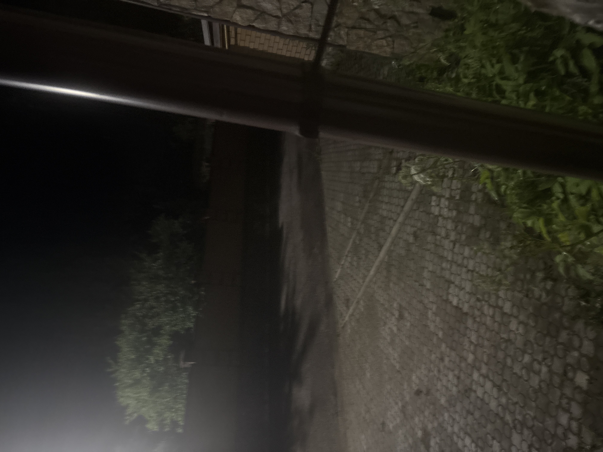 The image depicts an outdoor scene at night. In the foreground, a dark, cylindrical object, possibly a pipe or a pole, dominates the right side of the frame. To the left of this object is a cobblestone surface, and beyond that, a paved area with shadows cast upon it, suggesting artificial lighting. In the middle ground, a low, dark wall is visible, topped by dense, green foliage of trees or bushes, illuminated by a light source on the far left of the image. The background is a dark, indistinct expanse of night sky. The scene appears to be a residential or public area at night in the city of Ореховка, Russia.