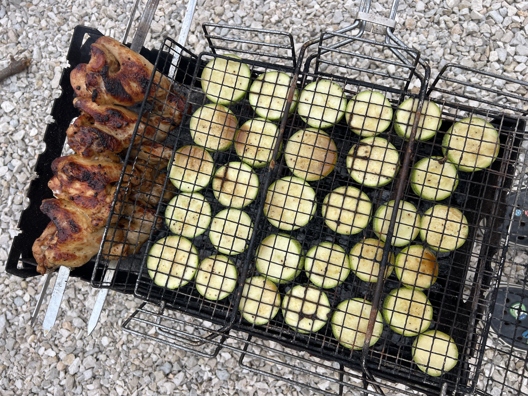 In an outdoor setting with a gravel ground, a grill basket is filled with food being cooked over charcoal. The top rack holds several pieces of grilled chicken, some with charred edges, skewered on metal rods. Below this, another grill basket contains numerous slices of zucchini, also showing grill marks and some browning. The scene appears to be an informal outdoor meal preparation.