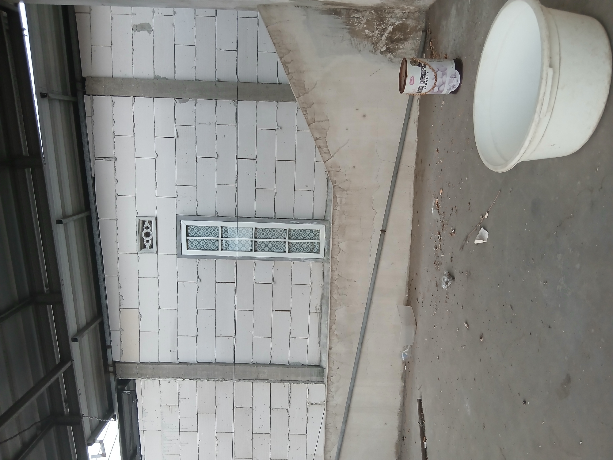 The image depicts an outdoor, unfinished construction area. A wall constructed of white cinder blocks is visible, with a single, decorative window. Two concrete pillars support an overhead metal roof structure. A sloping, cracked concrete wall runs along the lower portion of the scene. The ground is a concrete slab littered with debris, including cigarette butts and small pieces of paper. In the foreground, a large white plastic tub and a cylindrical container labeled "EGG DROPS Biskuit" are present. The overall impression is one of a site that is either under construction or has been abandoned. The location is identified as Kademangan Kulon, Indonesia.