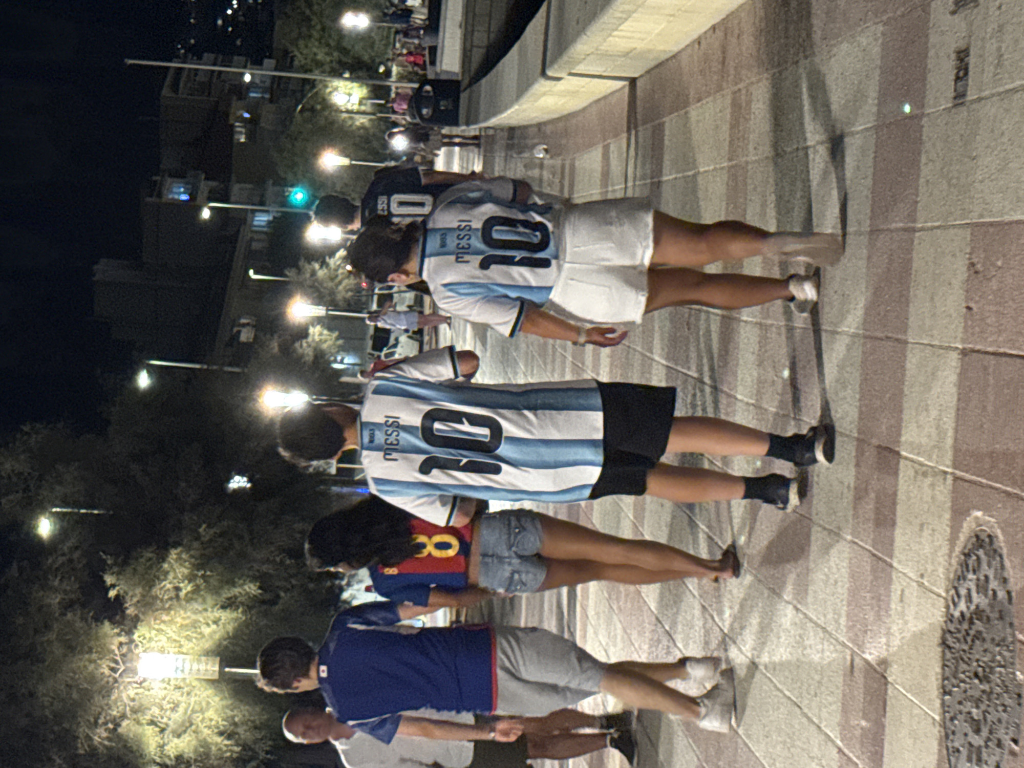 The image shows a group of people walking on a tiled promenade at night in Blanes, Spain. The promenade features a light-colored tiled surface with darker pinkish-brown rectangular accents. Streetlights with white light illuminate the area, casting shadows. In the background, there are trees with foliage and some buildings with lights.
The primary subjects are four individuals walking away from the camera.
The person furthest to the left is a man wearing a white shirt and dark shorts.
Next to him is a man wearing a blue soccer jersey with Japanese characters on the back and grey shorts.
To his right is a woman wearing a red soccer jersey with the number "8" and denim shorts.
Furthest to the right is a person wearing a white and blue striped soccer jersey with "MESSI 10" on the back and white shorts. The person to their left, further back in the frame, also appears to be wearing a similar soccer jersey.
Other individuals are visible further down the promenade, some appearing to be in small groups. A trash receptacle is visible on the right side of the promenade. The overall atmosphere suggests a casual evening outing in a coastal town.