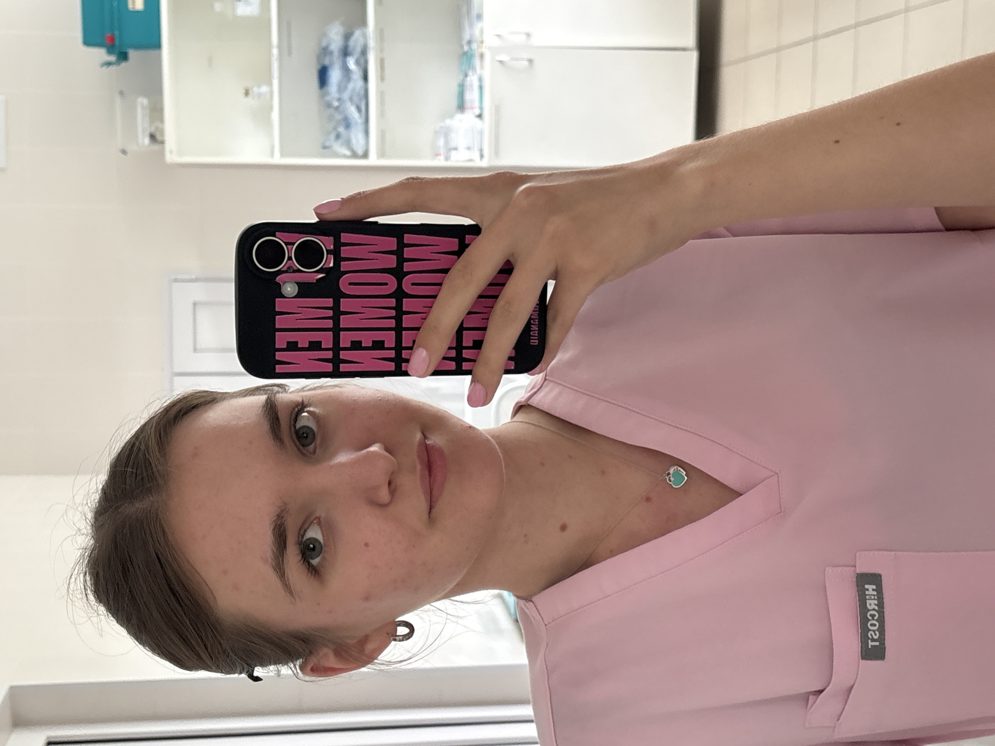 A fair-skinned person with light brown hair is taking a selfie. They are wearing a pink medical scrub with a necklace and earrings. The person is holding a black cell phone with the word "WOMEN" printed on the case in pink lettering. The background appears to be a sterile environment with white cabinets. The city is not discernible from the image.