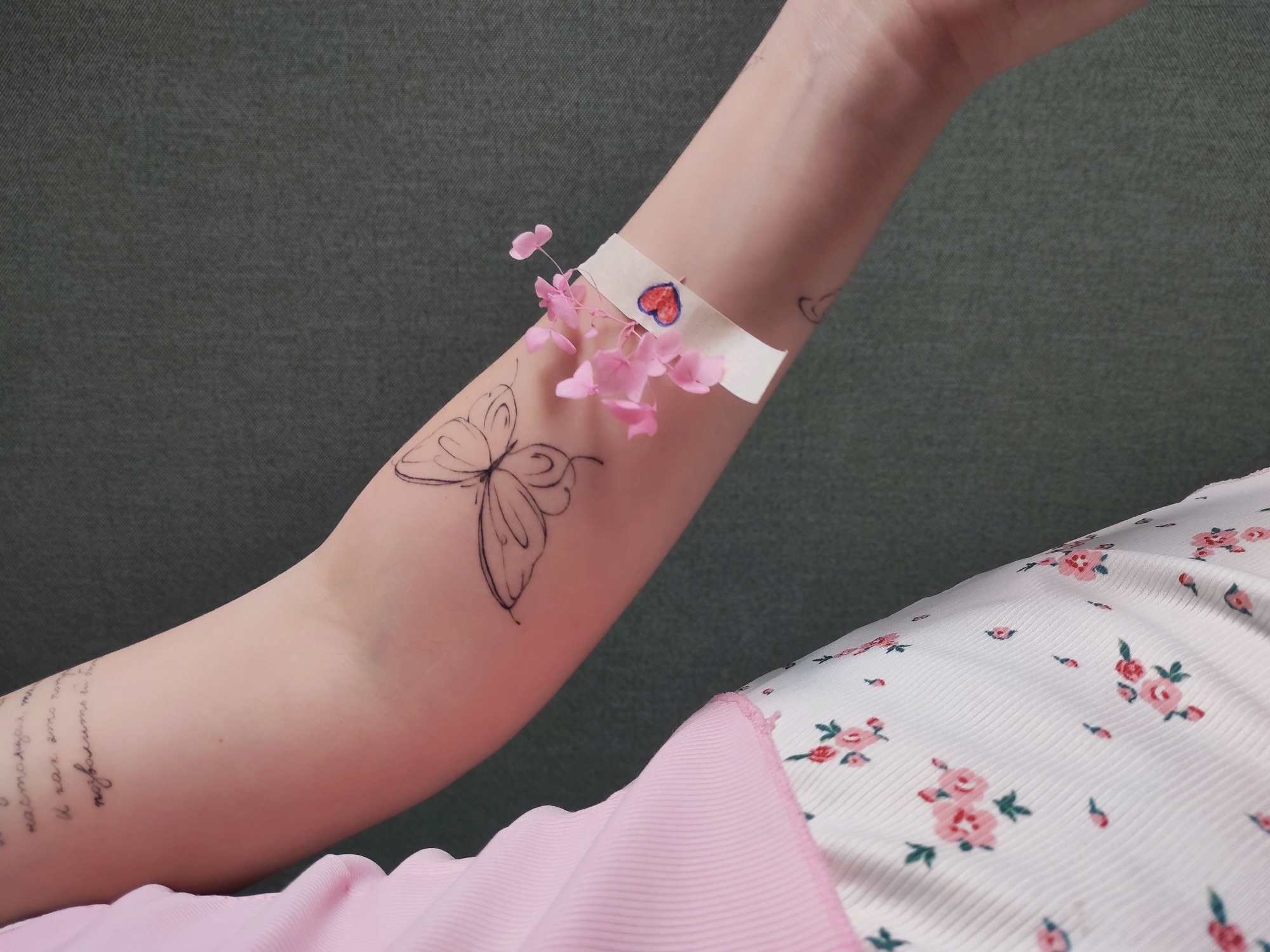 A person's forearm is shown with pink flowers affixed to the band-aid on the arm. The background is an olive green, and the dress is white with tiny pink flowers. The forearm has two tattoos, one a butterfly and the other words written in cursive. This image does not provide enough context to infer a location in Chaikovsky, Russia.