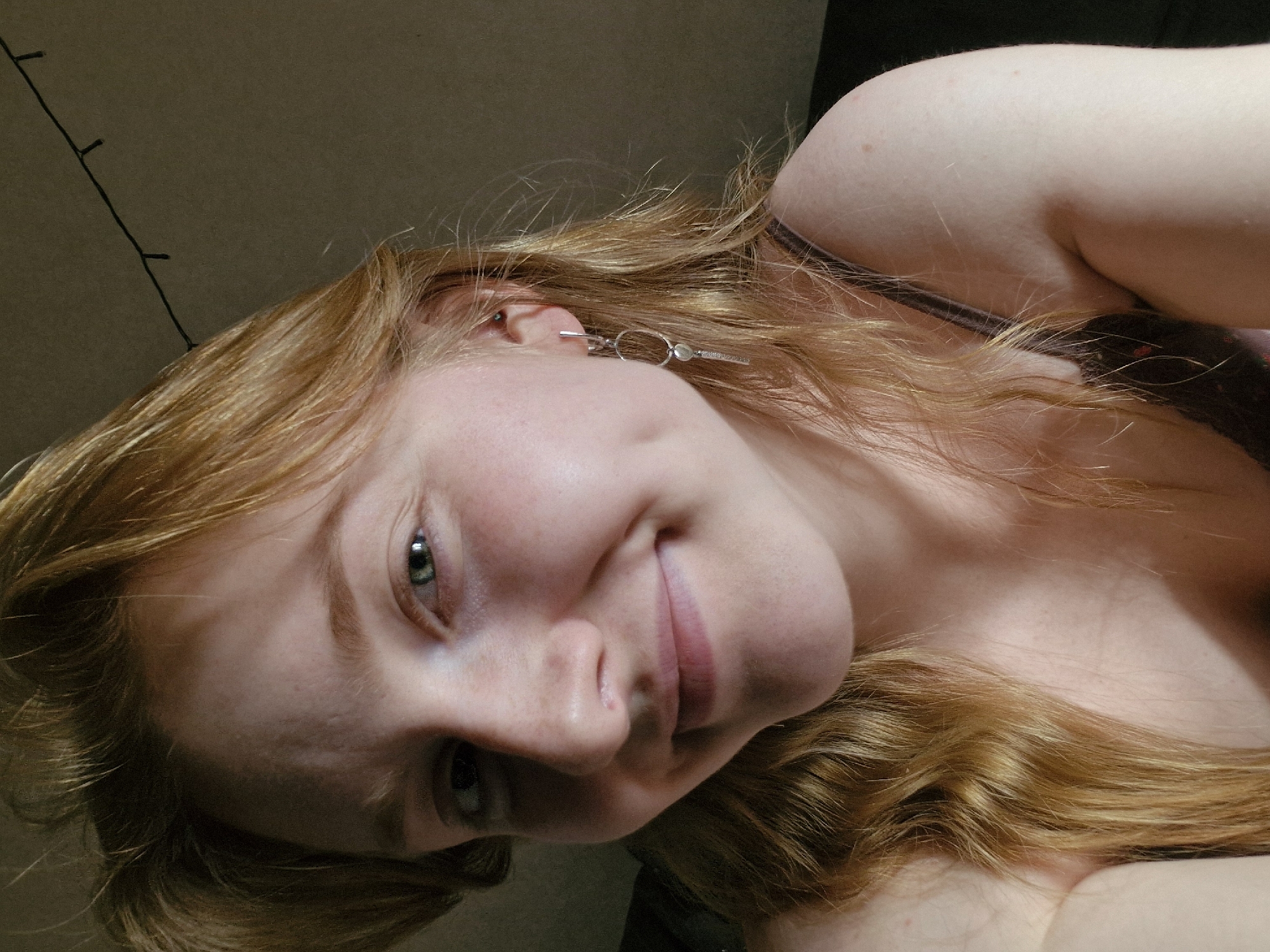 This close-up portrait captures a fair-skinned young person, likely female, with long, wavy, reddish-blonde hair, angled diagonally across the frame. The subject's head is tilted, and they gaze towards the viewer with their left eye, which appears to be green or hazel. A subtle smile or smirk is present on their lips, conveying a sense of contentment or slight playfulness.
Prominent facial features include a visible cheek with a hint of a dimple, part of the nose, and the lower lip. A silver-colored earring or decorative hair clip with a small hoop and a bar design is visible near the left ear, nestled within the flowing hair. A dark strap, possibly from a tank top or bra, is visible over the person's right shoulder.
The background is a plain, dark, and indistinct surface, suggesting an indoor setting. A thin, dark string of what appears to be decorative fairy lights runs diagonally across the upper left portion of the background. The lighting is soft and diffused, creating gentle shadows that highlight the contours of the face and hair.
The image portrays a personal moment, possibly taken in a relaxed indoor environment in Yekaterinburg, Russia. There is no visible text in the media file.