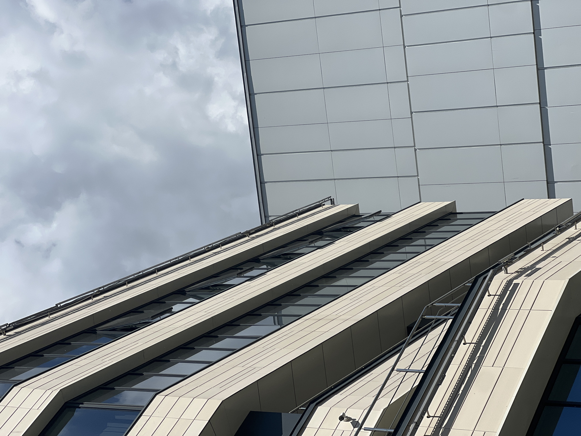 The image shows a close-up, low-angle view of a modern building with a grey, cloudy sky in the background. The building features a striking architectural design with multiple sections. One section, visible in the upper right, is clad in large, rectangular, light grey panels arranged in a grid. This section appears to be a flat surface extending upwards.
In the foreground and midground, a more complex section of the building is visible, characterized by angled, cream-colored cladding and long, horizontal windows that reflect the sky. These angled sections create a sense of dynamism and depth. The lines of the building are sharp and geometric. On one of the angled sections, a narrow metal structure, resembling a ladder or maintenance rail, runs diagonally. A security camera is also visible on this section. The overall impression is of a contemporary urban structure.
The time of day is difficult to ascertain precisely due to the overcast conditions, but the shadows suggest it is daytime. The weather appears to be cloudy or overcast, with a grey, diffused light. There is no text visible in the image. The location is Saint Petersburg, Russia, and the architecture is consistent with modern commercial or residential buildings found in major cities. No people are visible in the image.