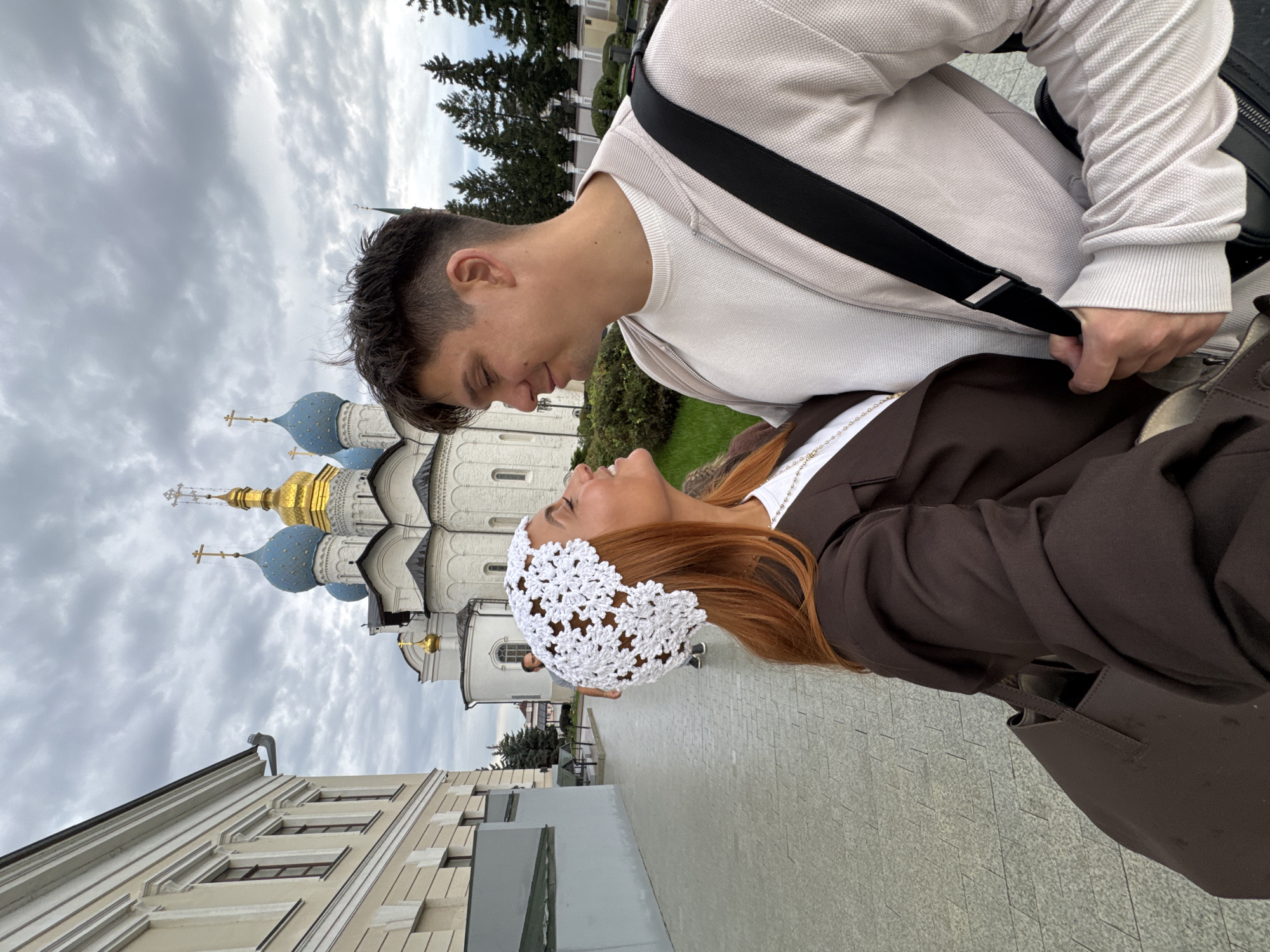 A young couple is captured in a close-up selfie in front of the Kazan Kremlin, a UNESCO World Heritage site in Kazan, Russia. The woman, with vibrant red hair, wears a white crochet headscarf and a brown blazer over a white top, adorned with a delicate gold chain necklace. The man, with dark, short hair, sports a light beige bomber jacket over a white t-shirt. They are gazing into each other's eyes, their faces nearly touching, conveying an intimate moment. In the background, the iconic Kul Sharif Mosque with its blue domes and golden spires is partially visible, set against a cloudy sky. The paved walkway and the facade of a historical building to the left of the couple suggest they are within the Kremlin grounds. Both individuals are carrying shoulder bags.
