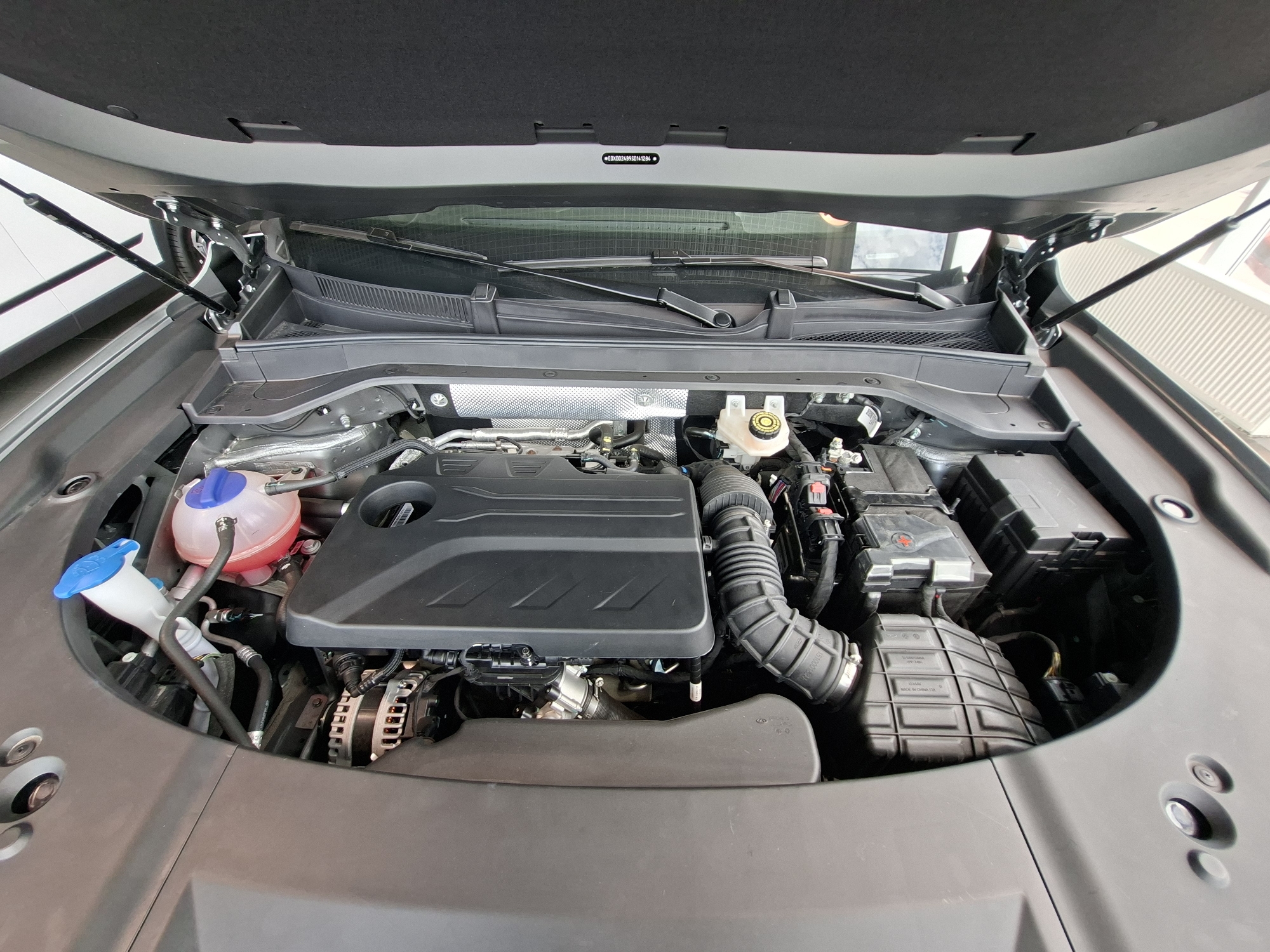 The image shows the engine bay of a vehicle, with the hood open. The engine components are visible, including the engine cover, air intake, alternator, and fluid reservoirs. The VIN sticker is visible on the underside of the hood. The scene appears to be indoors, possibly in a garage or showroom, with no visible indication of its specific location in Izhevsk, Russia. There are no people or actions depicted in the image. The environment is clean and well-lit.