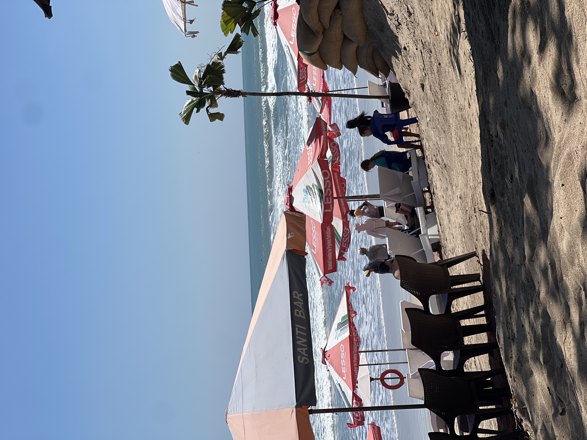 A bright, sunny day at Kuta Beach, Indonesia. The scene captures a section of the beach with several beach umbrellas, lounge chairs, and a few people. The prominent umbrella in the foreground has "SANTI BAR" printed on it. Other umbrellas display "LESSO Global Leader in Piping Solutions." Several sandbags are stacked to the right of the frame, possibly for erosion control. The ocean is visible in the background with moderate waves. Two children are seated on chairs in the midground, facing the ocean. A few adults are scattered near the umbrellas, some standing, some seated. The sand is light brown, and shadows from the umbrellas and vegetation are cast across it.