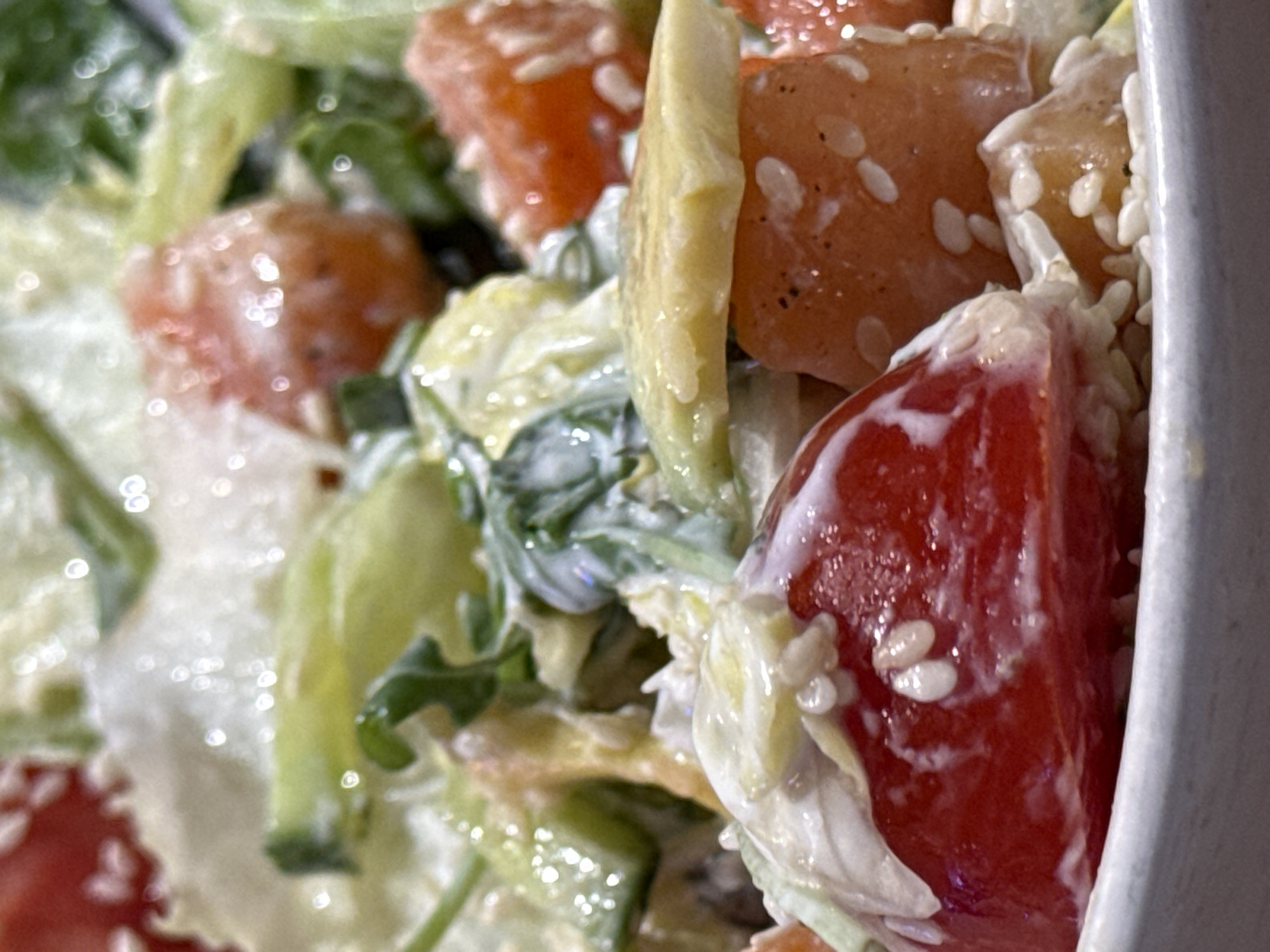 A close-up, overhead shot shows a salad in a white bowl. The salad consists of chopped tomatoes, cucumber, lettuce, and possibly avocado, all coated in a creamy dressing and sprinkled with sesame seeds. The lighting is bright, causing some specular highlights on the wet ingredients, particularly the tomatoes and dressing. The background is blurred and out of focus, indicating a shallow depth of field. This image was taken in Bar, Montenegro.