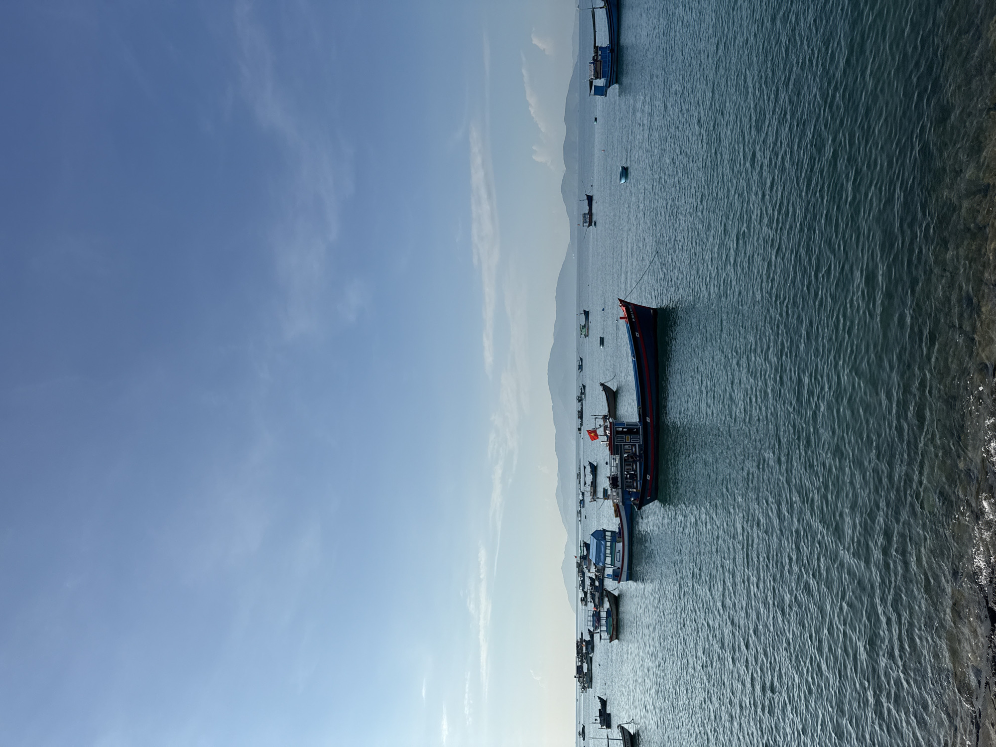 In Bac Nha Trang Ward, Vietnam, a body of water is filled with numerous fishing boats anchored under a clear blue sky with scattered white clouds. The boats vary in size and color, with some featuring red flags. In the background, a chain of mountains is visible. The foreground consists of the water's surface with gentle ripples and a rocky shore.