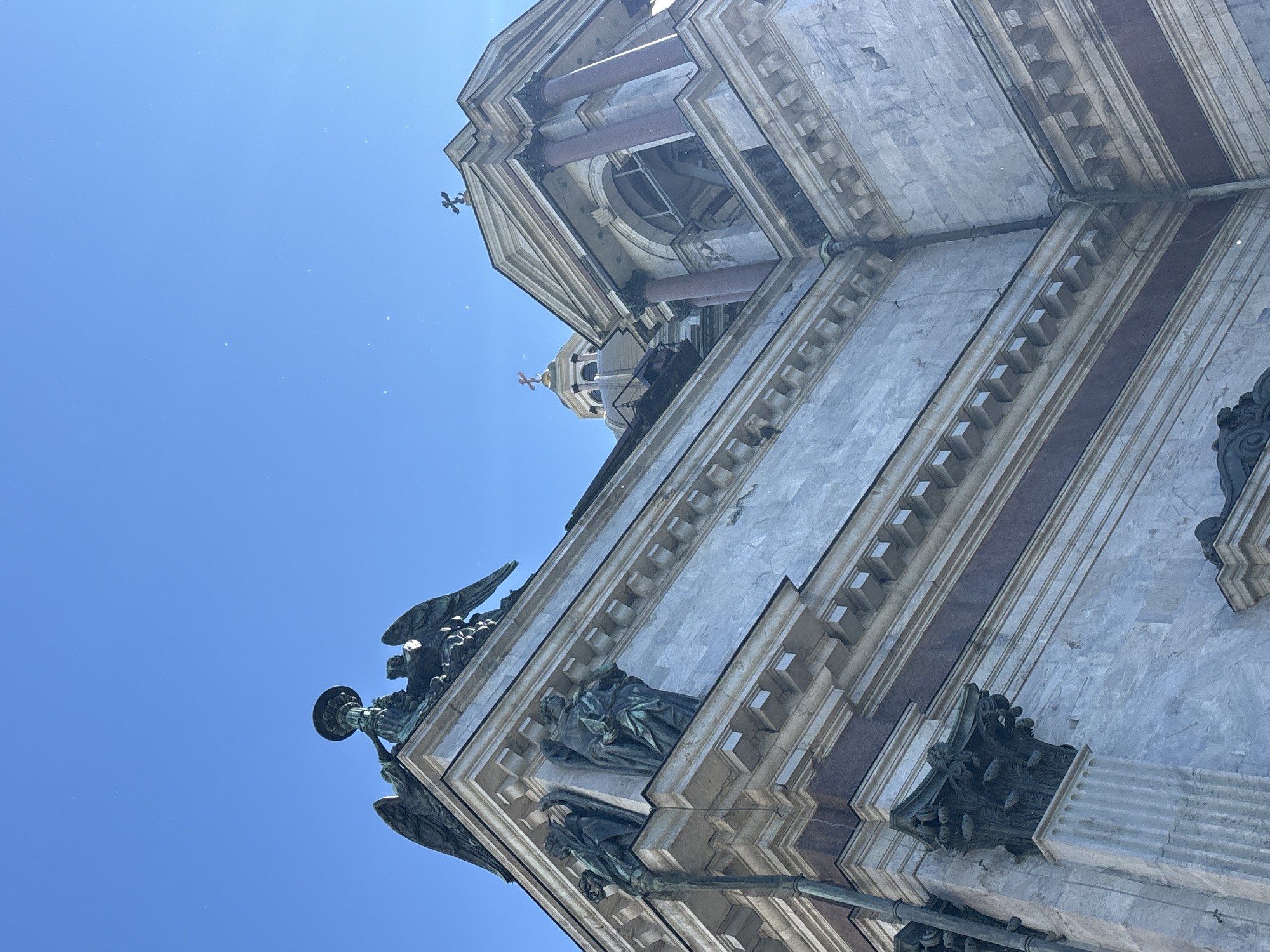 The image shows a low-angle view of the exterior of St. Isaac's Cathedral in Saint Petersburg, Russia, against a clear blue sky. The cathedral's facade is constructed of marble, with ornate architectural details, including columns, cornices, and sculptures. At the top of the building, bronze statues of angels are visible, one holding a candlestick. A cross is also visible atop a smaller dome in the background. The sun is shining, casting some shadows and highlighting the textures of the stone. The overall scene conveys a sense of grandeur and historical significance.