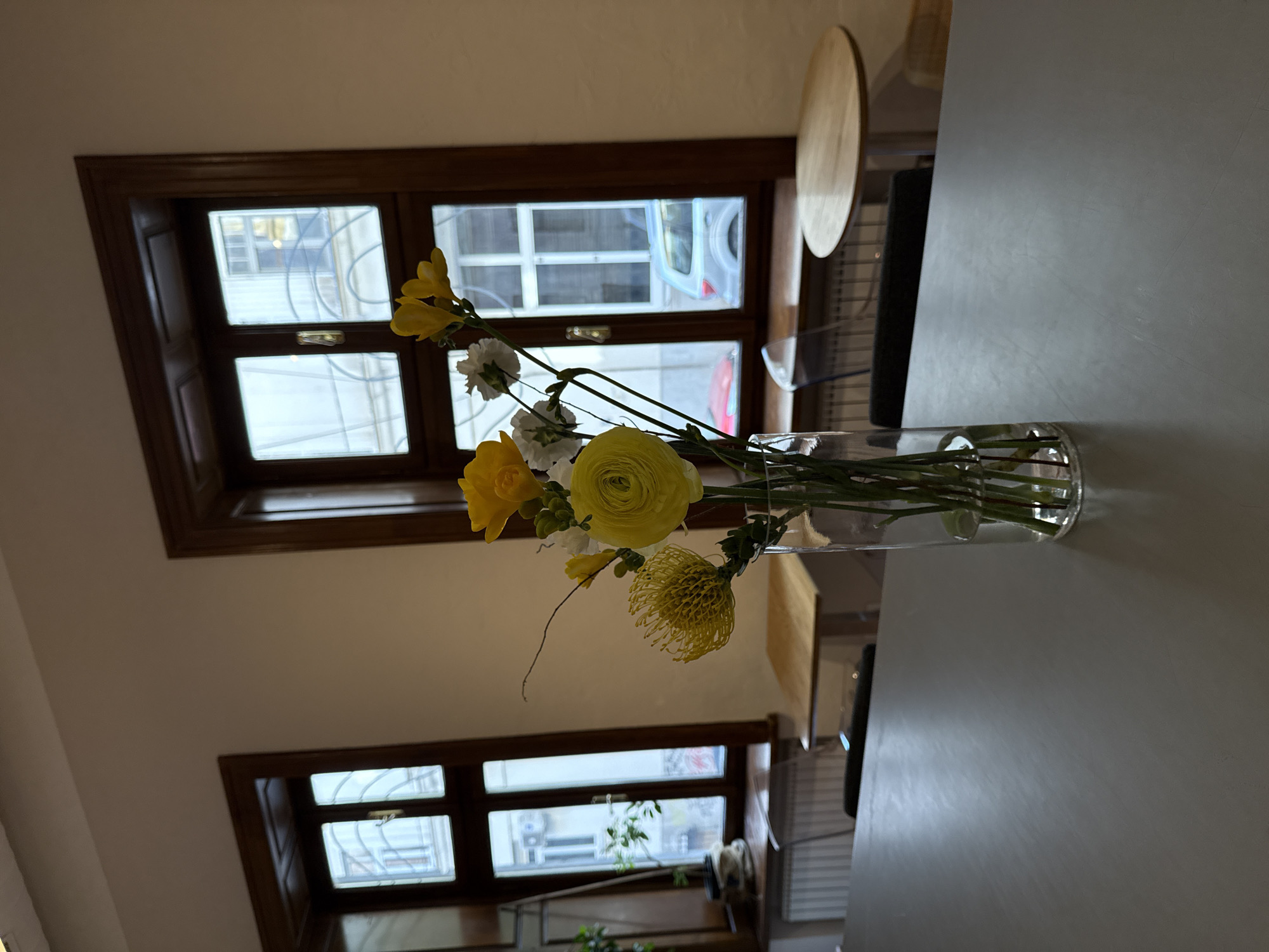 The image displays an indoor scene, likely a cafe or a well-decorated room, bathed in natural daylight. In the foreground, a transparent glass vase holds a vibrant bouquet of yellow and white flowers. Prominent yellow blooms include a tightly petaled ranunculus, several delicate freesias, and a spiky, globe-like flower. Smaller white flowers, possibly carnations, are interspersed among the green stems and leaves. The vase rests on a smooth, light grey tabletop.
In the background, two large windows with dark brown wooden frames provide a view of the outside. Through the upper window, glimpses of an adjacent building and what appears to be the door of a white vehicle are visible. The lower window reveals more outdoor structures and some sparse greenery, possibly from another plant. The walls of the room are a light, neutral color. A round, light-colored wooden object, possibly a decorative disc or part of a shelf, is partially visible on the right side of the upper background.
The lighting suggests it is daytime, with soft, diffused light entering through the windows. There is no discernible human activity or specific event taking place. The scene exudes a calm and aesthetically pleasing atmosphere. No visible text is present in the image. The location context provided is Belgrade, Serbia.