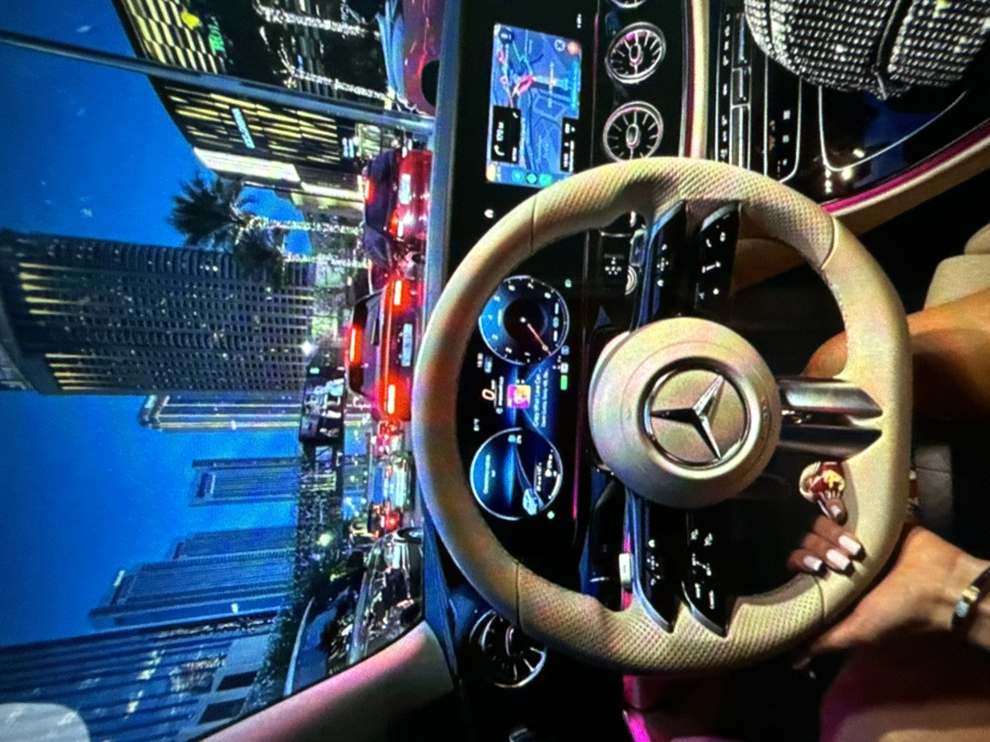 The image shows the interior of a car, likely a Mercedes-Benz, with a focus on the steering wheel. A person's hands are visible on the steering wheel, and the dashboard features screens displaying navigation and vehicle information. Outside the car's window, a cityscape is visible with tall buildings, suggesting an urban environment. The city appears to be Unknown, Unknown. There are other vehicles and traffic visible on the screen suggesting some form of simulation. The overall scene appears to be taken inside the car.
