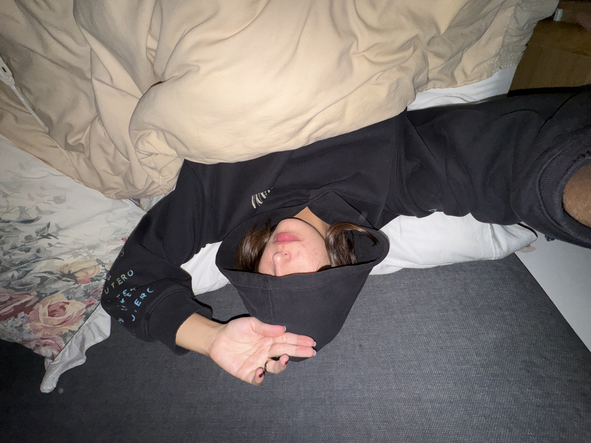 The image shows a person lying in bed under a beige comforter. The person is wearing a black hooded sweatshirt, with the hood pulled over their head. Their face is partially visible, and one hand is resting on the gray bedding. The location is indeterminate from the image.