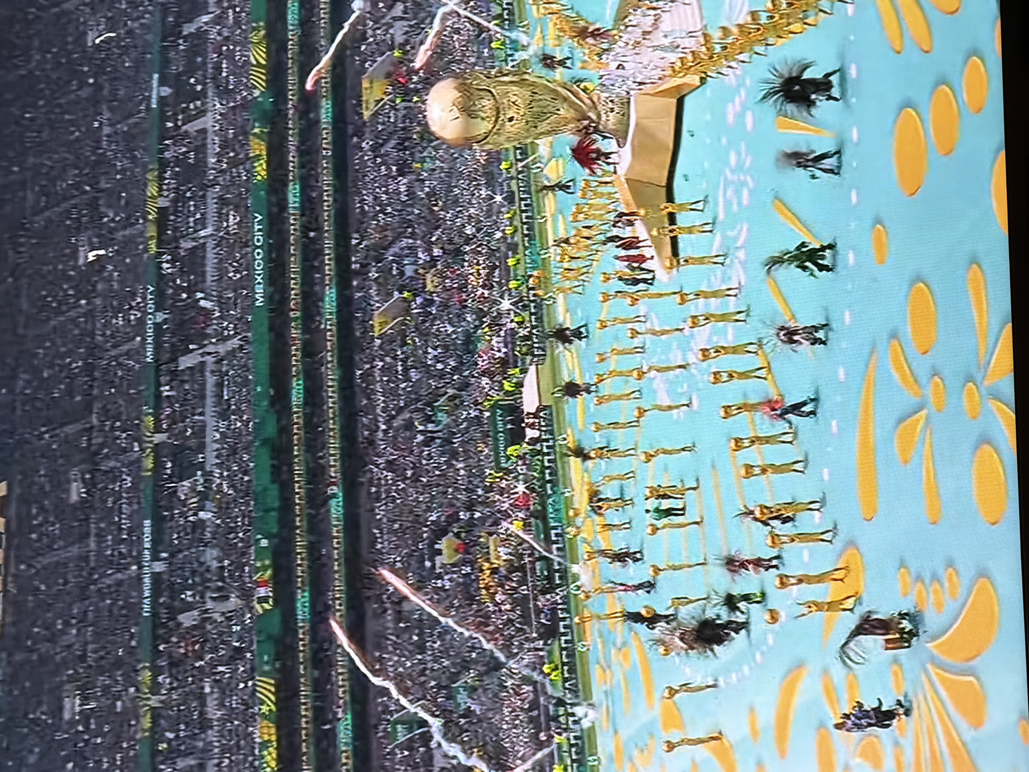 The image depicts a scene from a stadium during an event, likely an opening ceremony for a major sporting competition. In the background, a large crowd fills the stadium seating. In the foreground, performers are spread across a light blue surface decorated with yellow floral patterns. Many performers are dressed in gold costumes and appear to be holding golden spheres above their heads. A large, prominent golden trophy, resembling the FIFA World Cup trophy, is situated on a raised platform towards the right side of the image. There are also several performers in more elaborate, dark costumes with feathered headdresses. Fireworks or pyrotechnics are visible in the upper left and right portions of the frame, with streaks of light arcing across the sky. Signs in the stadium seating indicate "MEXICO CITY" and "FIFA WORLD CUP RUSSIA." The lighting suggests it is nighttime or an indoor venue with artificial lighting. There is no indication of the city of Pazardzhik, Bulgaria.