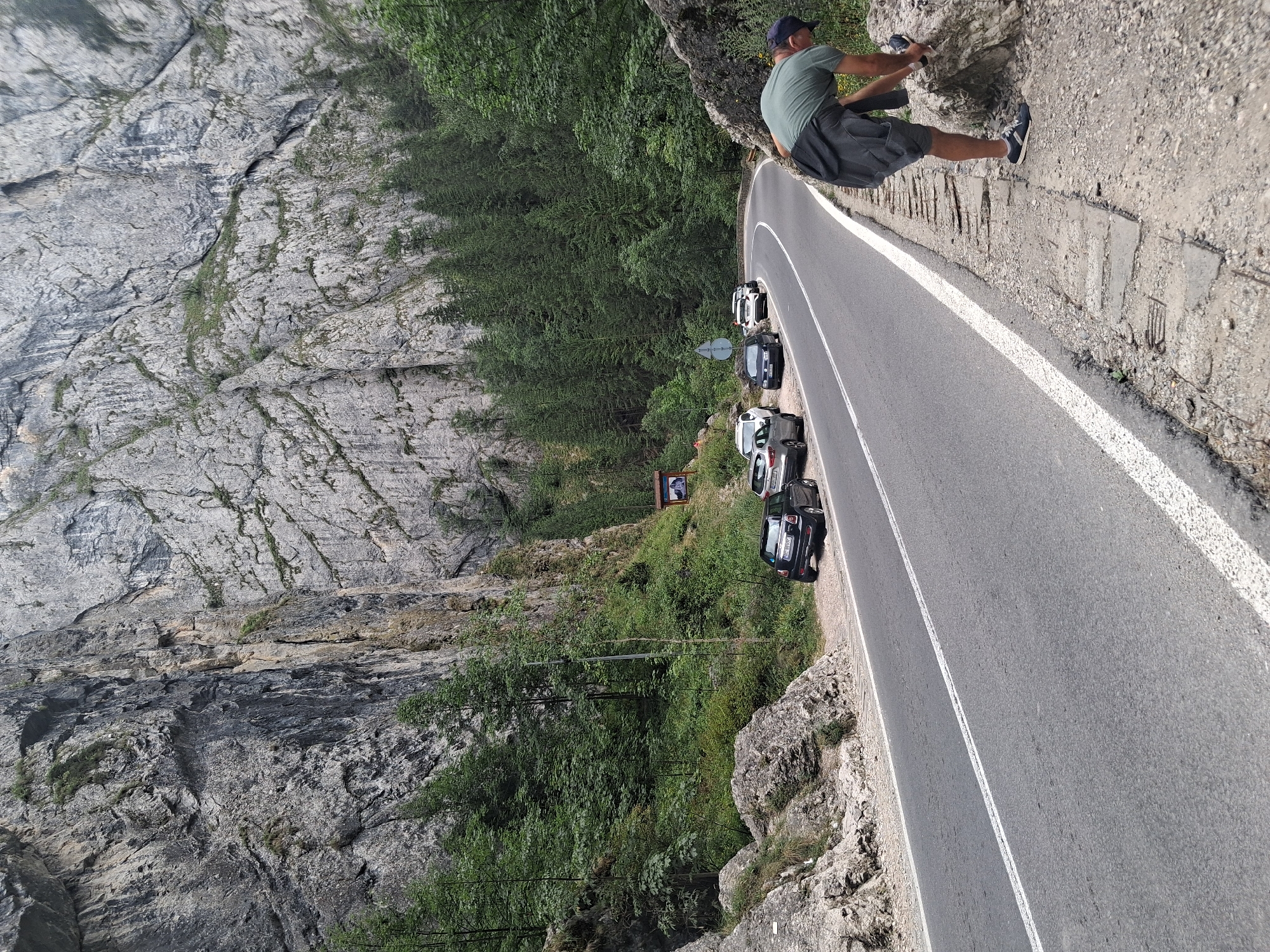 The image depicts a scenic road winding through a mountainous area with a towering, sheer rock face on the left side and dense green forest on the right. Several cars are parked along the right side of the road, suggesting a popular scenic spot or trailhead. In the foreground on the right, a person wearing a green t-shirt, shorts, and a baseball cap is bent over, appearing to interact with a rock. The road is paved with white lane markings and a white shoulder line. A brown sign is visible further up the road, indicating a point of interest. The overall environment is natural and rugged, characteristic of a mountainous region. This scene is located in Bicaz-Chei, Romania.
