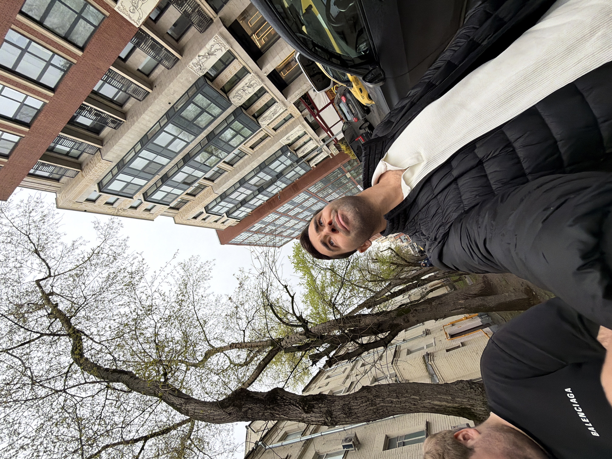 The image depicts a low-angle perspective of an individual taking a selfie in an urban environment.
People: Two men are visible in the frame. One man, presumably taking the photo, is wearing a white ribbed sweater and a black puffer jacket. The second man, only partially in frame, is wearing a black t-shirt with the word "BALENCIAGA" printed on it.
Subjects: The primary subject is the person taking the selfie and the surrounding environment.
Objects: Buildings, trees, and possibly vehicles are present in the background. A yellow taxi is parked on the street.
Actions: The person taking the selfie is holding a phone or camera.
Interactions: The two men appear to be together, possibly interacting.
Environment: The location is an urban setting, likely a street in Moscow, Russia. This can be deduced from the architectural style of the buildings, which are common in Moscow. The weather appears to be overcast, based on the sky. Trees are partially bare, indicating it is likely early spring.