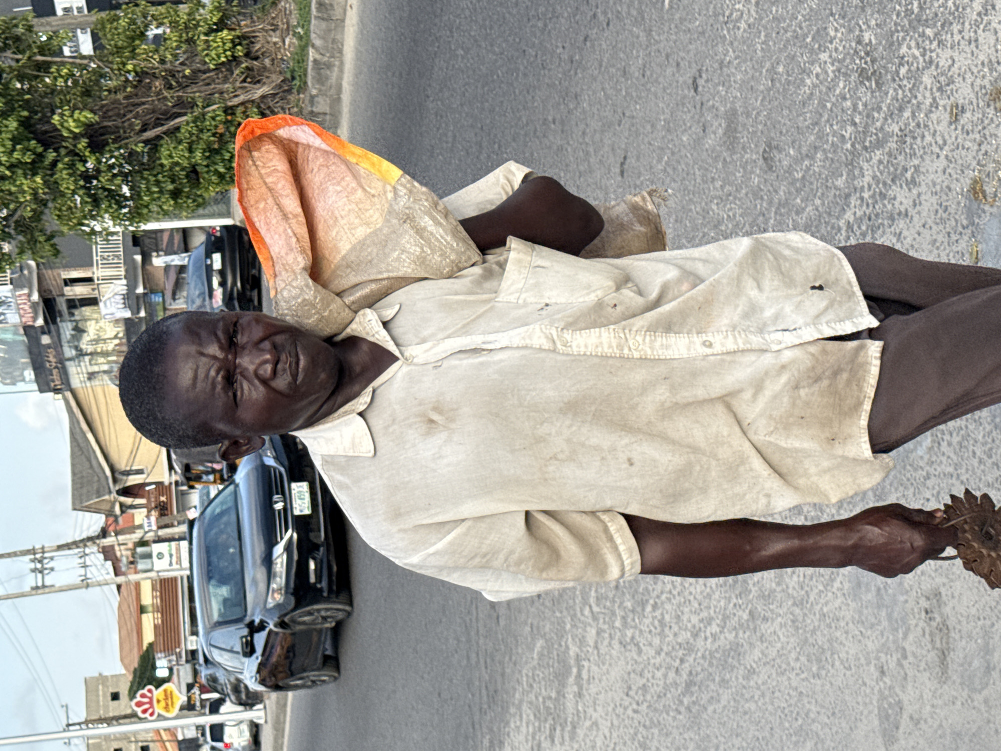 In Ado, Nigeria, a dark-skinned man with a deeply lined face is walking on a city street. He carries a bag over his shoulder and something indistinct in his right hand. He wears a light-colored, button-down shirt. Behind him are cars, buildings, power lines, and trees, indicating an urban environment.