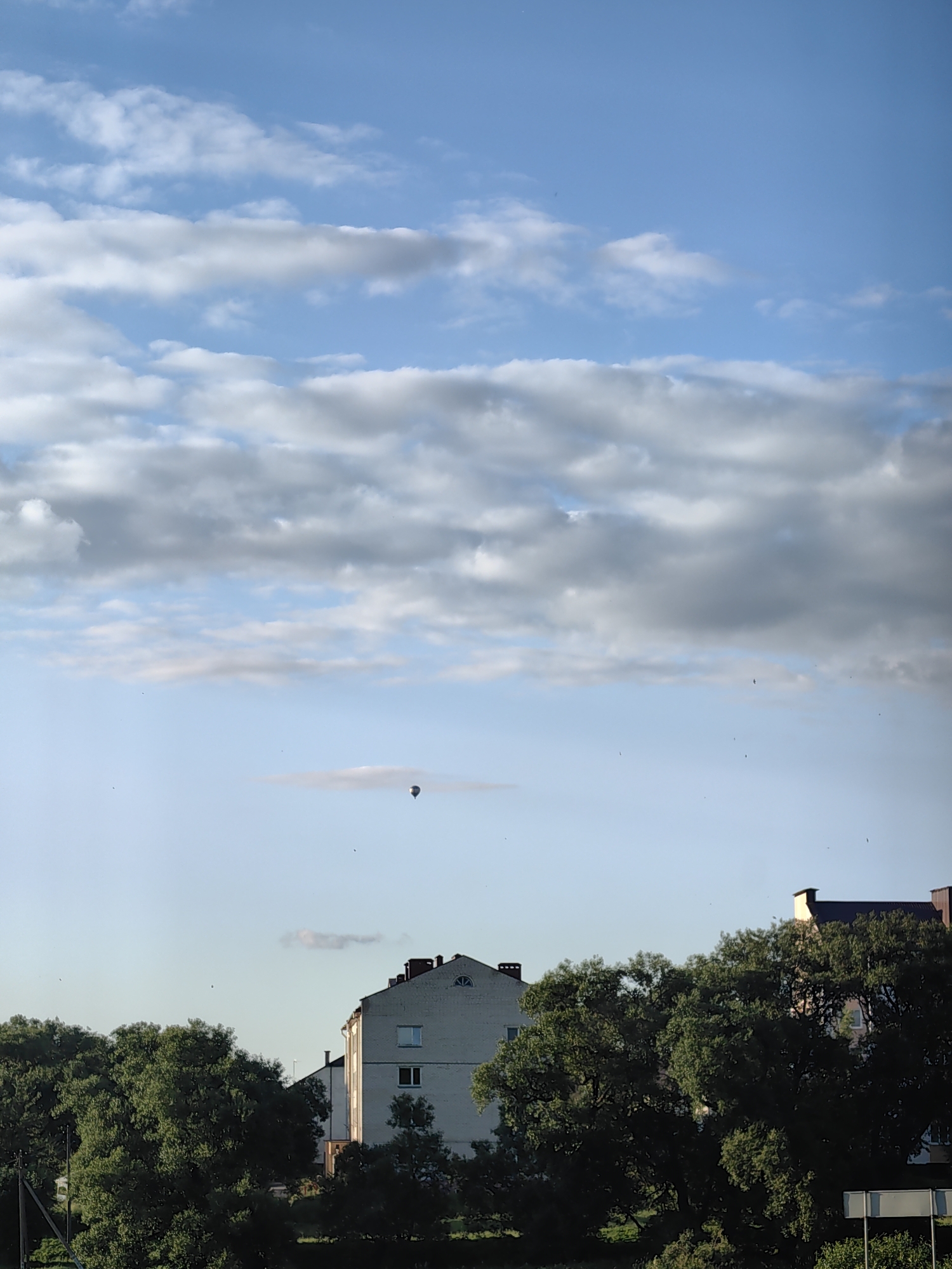 In the city of Fanipolski Sel'ski Savet, Belarus, a clear sky with scattered white and gray clouds dominates the upper portion of the image. A hot air balloon is visible in the middle distance, appearing as a small dark shape against the pale blue sky. Below the balloon, a thin, elongated cloud stretches horizontally. In the foreground, a light-colored, multi-story building with visible windows and a peaked roof stands partially obscured by lush green trees. To the right of this building, another building with a reddish-brown roof is also visible. The lower part of the image is filled with dense foliage from large trees, with some smaller trees and vegetation visible at the very bottom. The scene appears to be captured during daylight, possibly in the late afternoon or early evening, given the quality of light on the buildings.