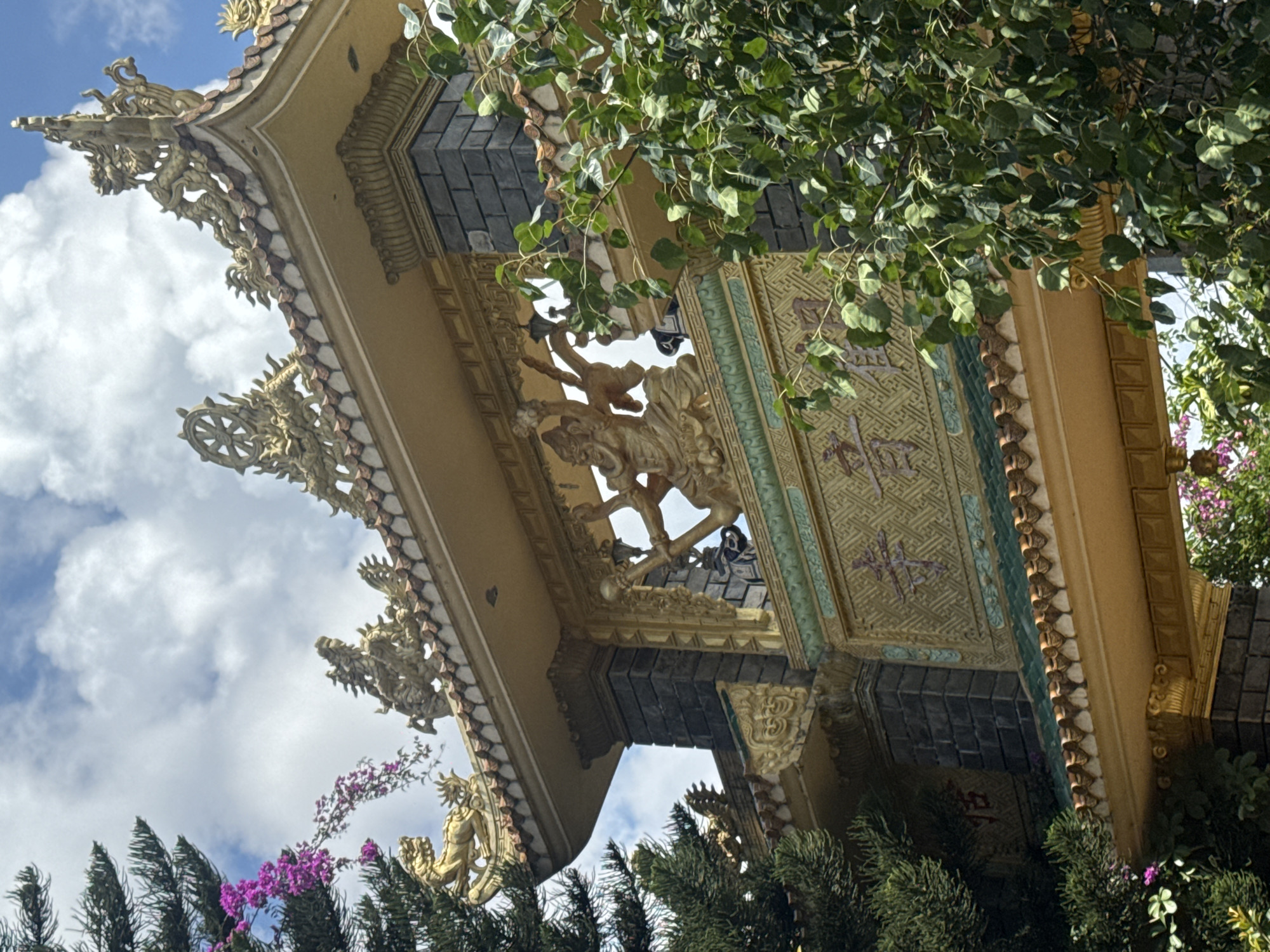 The image shows the ornate entrance gate of a Buddhist temple, likely Linh Phuoc Pagoda, in Da Lat, Vietnam. The gate is constructed with dark brick pillars and topped with a gilded, multi-tiered roof adorned with dragon figurines and Buddhist motifs like the Dharma wheel. A central archway reveals a golden statue of a multi-armed deity. The background features a bright blue sky with scattered white clouds, partially obscured by lush green foliage and a sprig of vibrant pink bougainvillea. The overall scene depicts a serene and culturally rich architectural landmark within a natural setting.