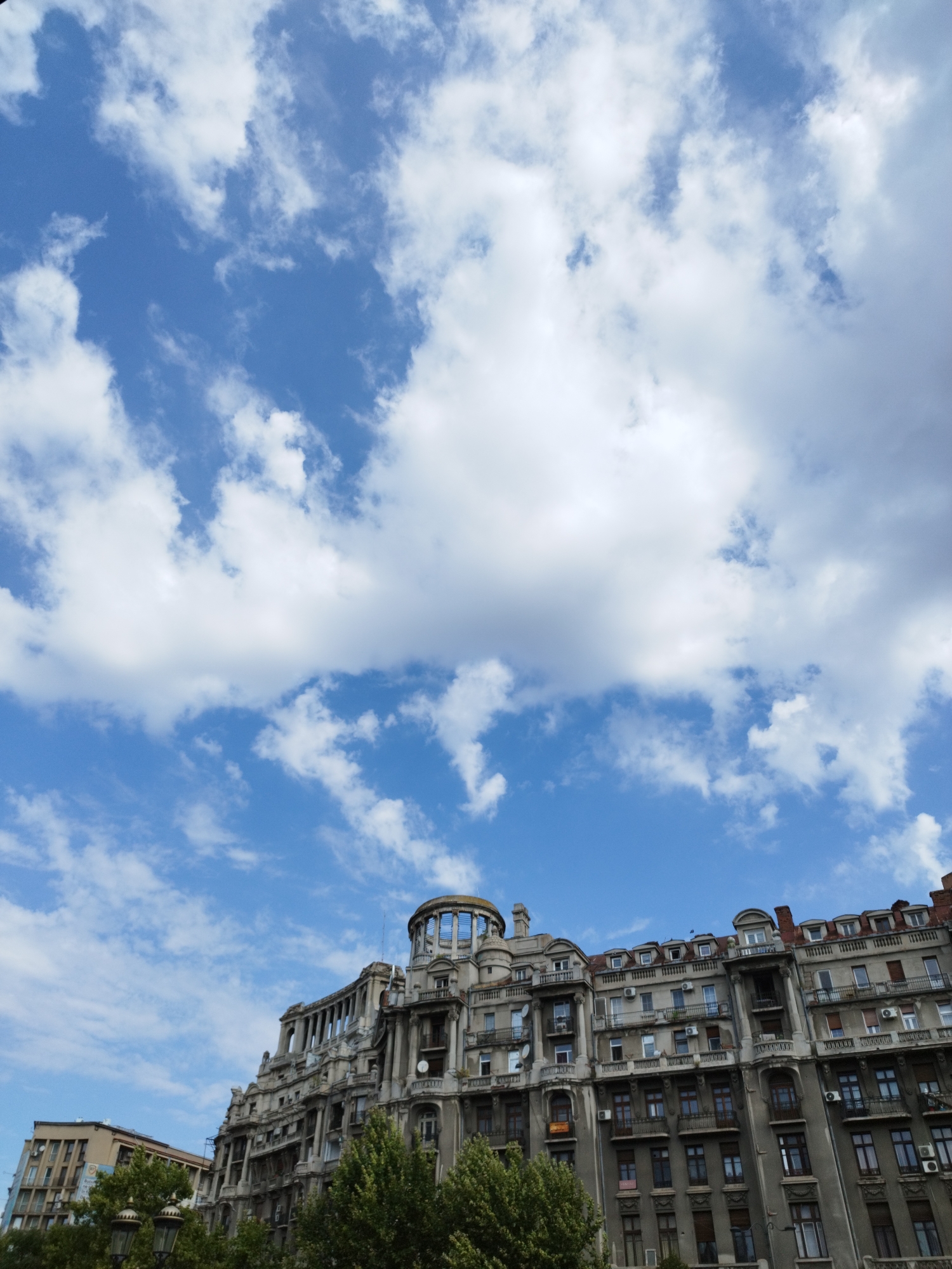 The image shows an ornate, historical building in Bucharest, Romania, with a bright blue sky and white clouds filling the upper portion of the frame. The building appears to be a grand, multi-story structure with intricate architectural details, including balconies, columns, and a domed cupola at the top. The facade of the building is a neutral grey color, and there are many windows visible. Trees are in the foreground, obscuring the lower parts of the building and the street level. The sky is dynamic, with a mixture of large cumulus clouds and smaller, wispy clouds. The overall impression is that of an urban landscape with a focus on architectural grandeur under a lively sky.