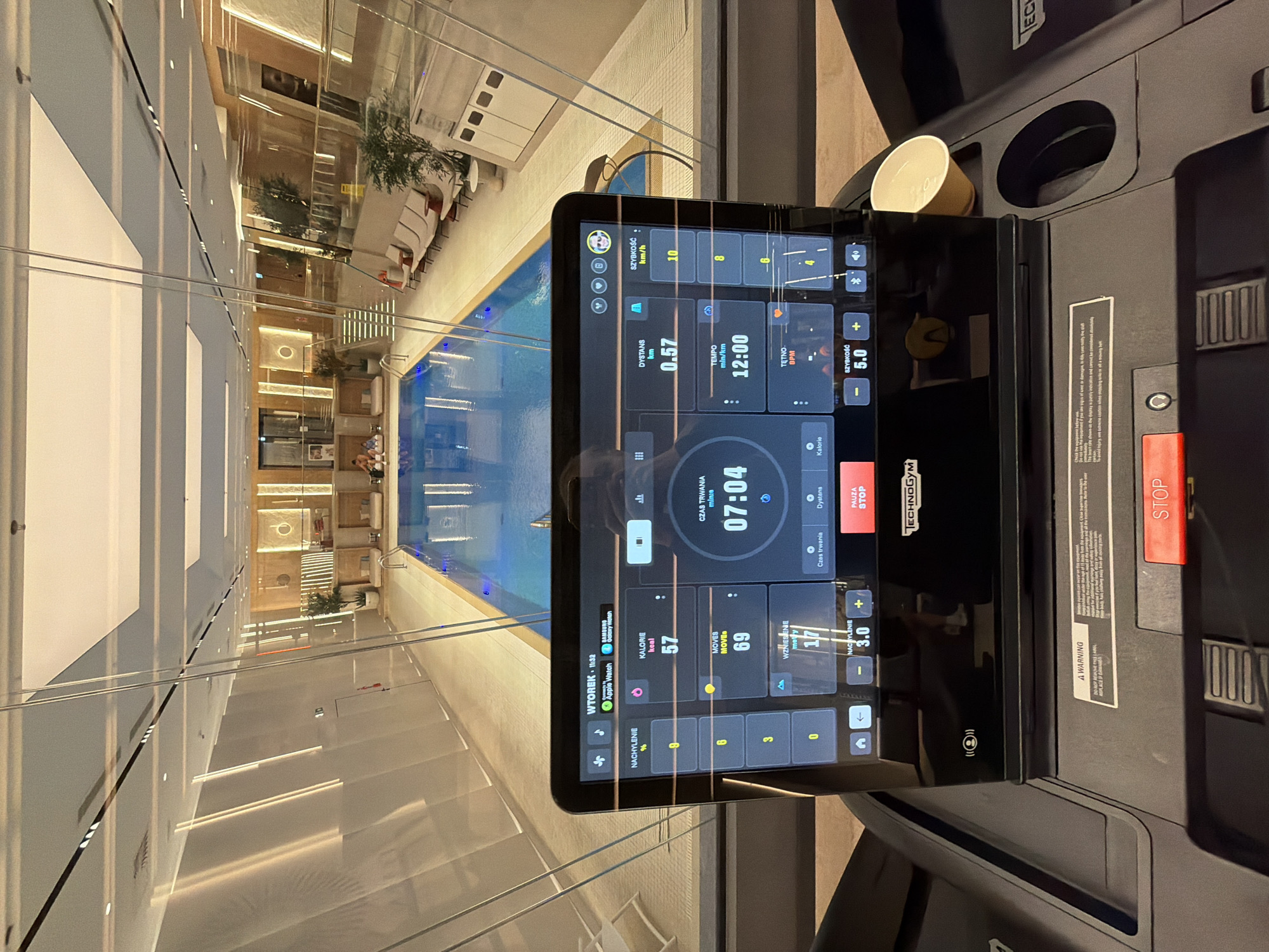 Here's a detailed description of the image:
**Scene:**
The image is taken from the perspective of someone on a Technogym treadmill, which is positioned indoors near a glass wall or balcony. The primary view through the glass is a luxurious interior space, likely within a building in an unidentified city.
**Environment:**
* The interior space beyond the glass is upscale, featuring a large indoor swimming pool.
* There are lounge chairs and potted plants arranged around the pool area, suggesting a resort or high-end residential setting.
* The architecture is modern with clean lines and a neutral color palette.
* The treadmill's screen shows fitness metrics such as time elapsed (07:04), distance (0.57 km), speed (10 km/h), calories burned (57 kcal), and other statistics.
**Objects:**
* **Treadmill:** A Technogym treadmill is the central object in the foreground. Its console is visible, displaying workout data.
* **Screen:** The treadmill's screen shows information about the exercise session, including duration, distance, speed, calorie burn, and other workout metrics.
* **Swimming Pool:** Visible through the glass wall is a large, well-lit indoor swimming pool.
* **Furniture:** Lounge chairs and potted plants are seen around the pool area.
**Overall Impression:**
The image conveys a sense of luxury and fitness. The location appears to be an exclusive spa or resort, where one can exercise while overlooking a stylish pool area. The treadmill data suggests the person has been exercising for a short period.