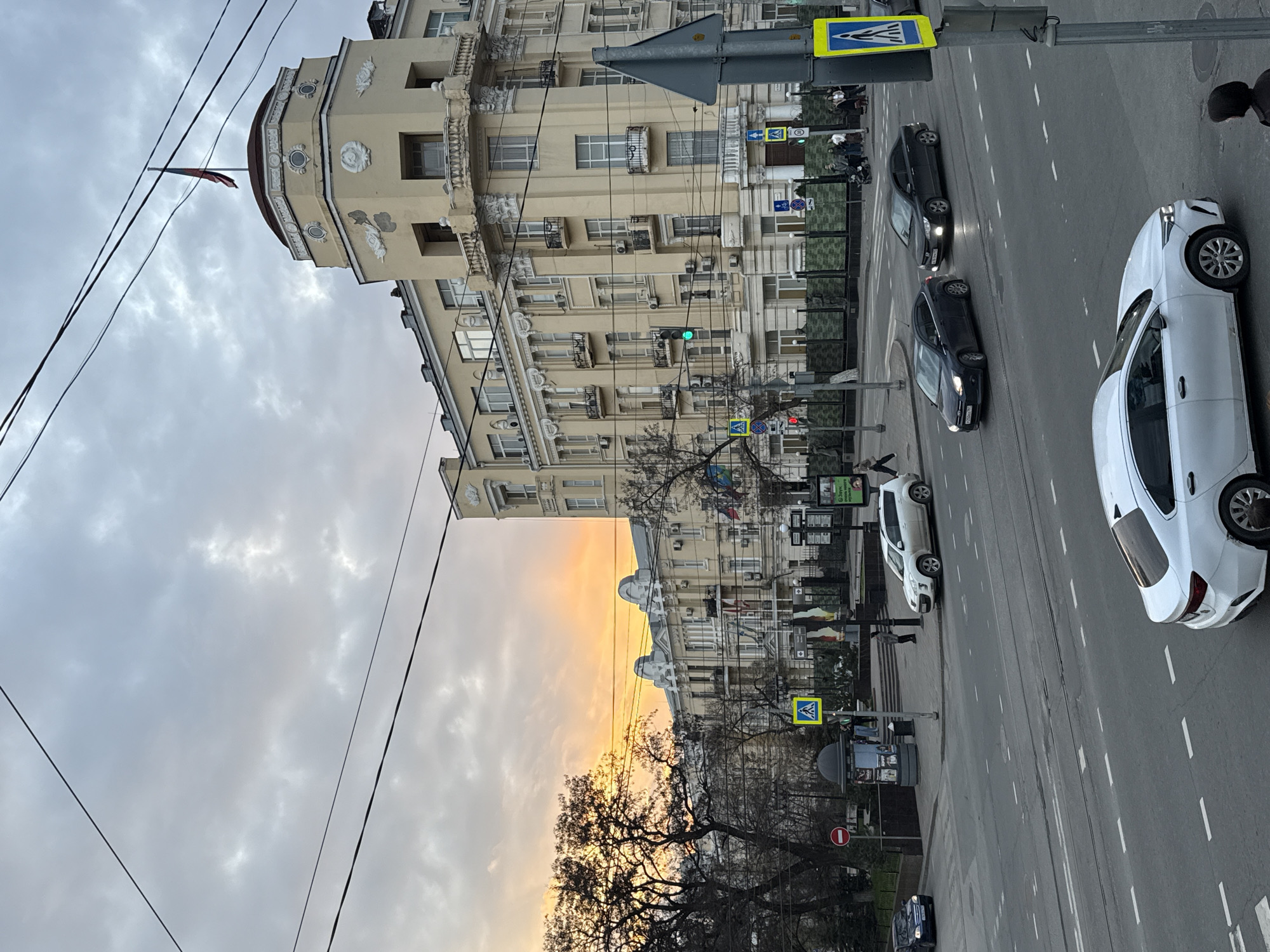 The image depicts a street scene in Rostov-on-Don, Russia, with a view looking towards the east during a sunset. A multi-story building with classical architectural elements occupies the left side of the frame, with a red flag visible atop its dome. Overhead power lines are strung across the sky. Below, trees line the street, obscuring the base of the building. In the foreground, a pedestrian crossing with signs marks the road. Three cars are visible on the street: a white sedan and two black sedans. A green traffic light is visible. A few people can be seen in the background. The sky is cloudy with hues of orange and gray reflecting the setting sun.