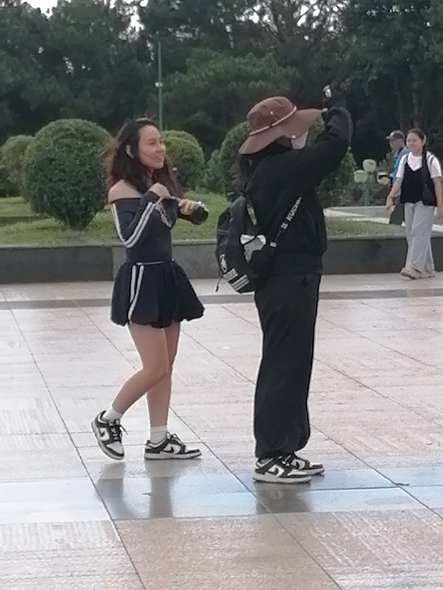 In Da Lat, Vietnam, two young women are captured mid-action on a paved plaza surrounded by lush green trees and manicured bushes. The woman on the left, sporting a dark, off-the-shoulder dress with white stripes and a flared skirt, is looking to her right with a smile and appears to be holding a small object, possibly a camera or phone. She is wearing white socks and black and white sneakers. The woman on the right, dressed in all black with a brown hat and a backpack, is facing away from the camera and appears to be taking a photograph with a phone held in her right hand. Her black pants are loose-fitting, and she is wearing black and white sneakers. In the background, another woman in a white shirt and black vest, with light-colored pants, walks away from the viewer. The overall scene is bright, with shadows indicating it is daytime.