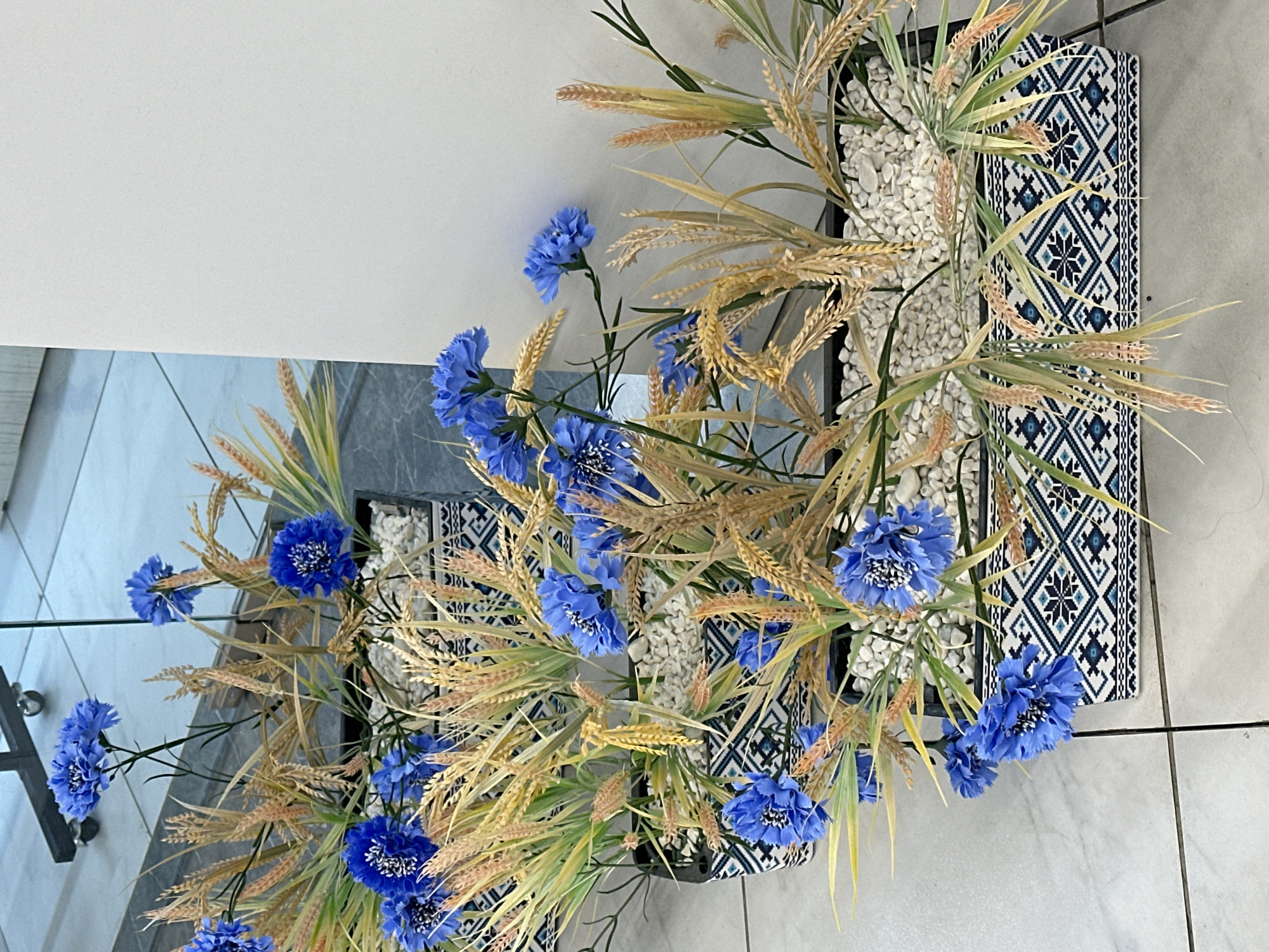 An indoor floral arrangement is presented in a bright setting. The primary subjects are numerous artificial blue flowers, resembling cornflowers with frilly petals and lighter centers, intermingled with artificial golden-brown and light green wheat stalks or ornamental grasses.
These elements are arranged densely within dark, rectangular planters that are filled with small white decorative pebbles. The visible sides of the planters feature a striking geometric pattern in shades of dark blue, light blue, and white, composed of squares and diamond shapes, reminiscent of traditional or ethnic embroidery.
The planters are positioned on a light grey tiled floor, characterized by visible grout lines. In the background, there is a plain, light-colored wall or surface. To the left of the arrangement, a reflective surface, possibly glass, shows a subtle and indistinct dark reflection.
No specific activity or event is taking place, as it is a static display. The lighting suggests an interior, possibly during the daytime. No explicit text is clearly visible within the image.