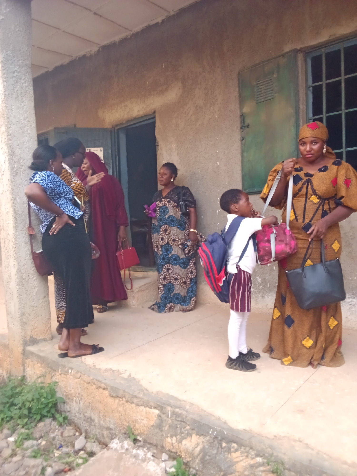 In Kakuri Gwari, Nigeria, a group of five individuals, consisting of four adult women and one child, are gathered on a concrete patio outside a building with a stucco exterior. The building features a large, green metal door and a barred window. The patio is slightly elevated from the ground, which is covered with dirt and vegetation.
On the left, three women stand near a pillar. The woman closest to the pillar wears a blue and white patterned top and a long black skirt, with a brown bag slung over her shoulder. Next to her, a woman in a yellow and black patterned dress is talking to another woman who is wearing a long, burgundy robe and a matching headscarf. The woman in the burgundy robe is holding a red handbag.
To the right, a child wearing a white shirt, striped pants, and white leggings stands between two women. The child has a blue and red backpack on their back and is holding a pink and red bag. The woman to the child's right wears a long, mustard-yellow dress with a diamond pattern and a matching headscarf, carrying a black tote bag in her left hand and a mobile phone in her right. Another woman in a patterned blue and brown dress is visible further to the right, holding a purple flower. The women appear to be engaged in conversation, with varying levels of interaction.
