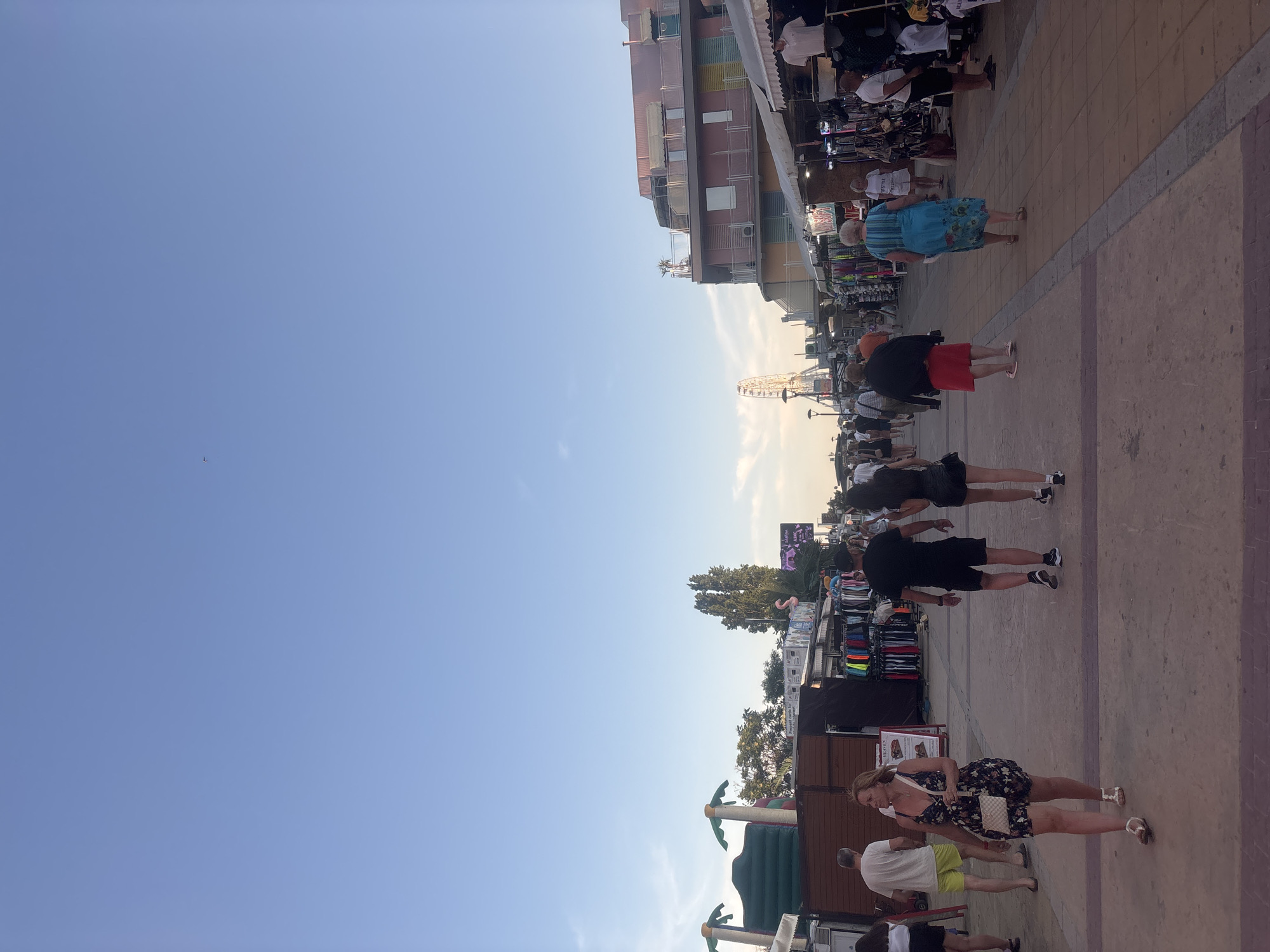 The image depicts a street scene in Nesebar, Bulgaria, likely during the evening as indicated by the lighting. A pathway, paved with reddish-brown tiles and bordered by grey stone, leads into the distance. The pathway is lined on both sides with market stalls selling various goods, including clothing. Several people are walking along the pathway, some individually and others in groups.
On the left side of the pathway, market stalls are visible, some featuring inflatable palm trees and what appears to be a bouncy castle. A man and woman are walking away from the camera, with the woman wearing a floral dress and carrying a handbag. Further down the path, more stalls and people can be observed.
On the right side of the pathway, market stalls display clothing items. A pink building with multiple windows and balconies is situated behind these stalls. Further in the distance, a Ferris wheel is visible against the sky. The sky itself is a pale blue, with a few wispy clouds and a small, distant object that could be a bird or a small aircraft. The overall atmosphere suggests a lively tourist area.