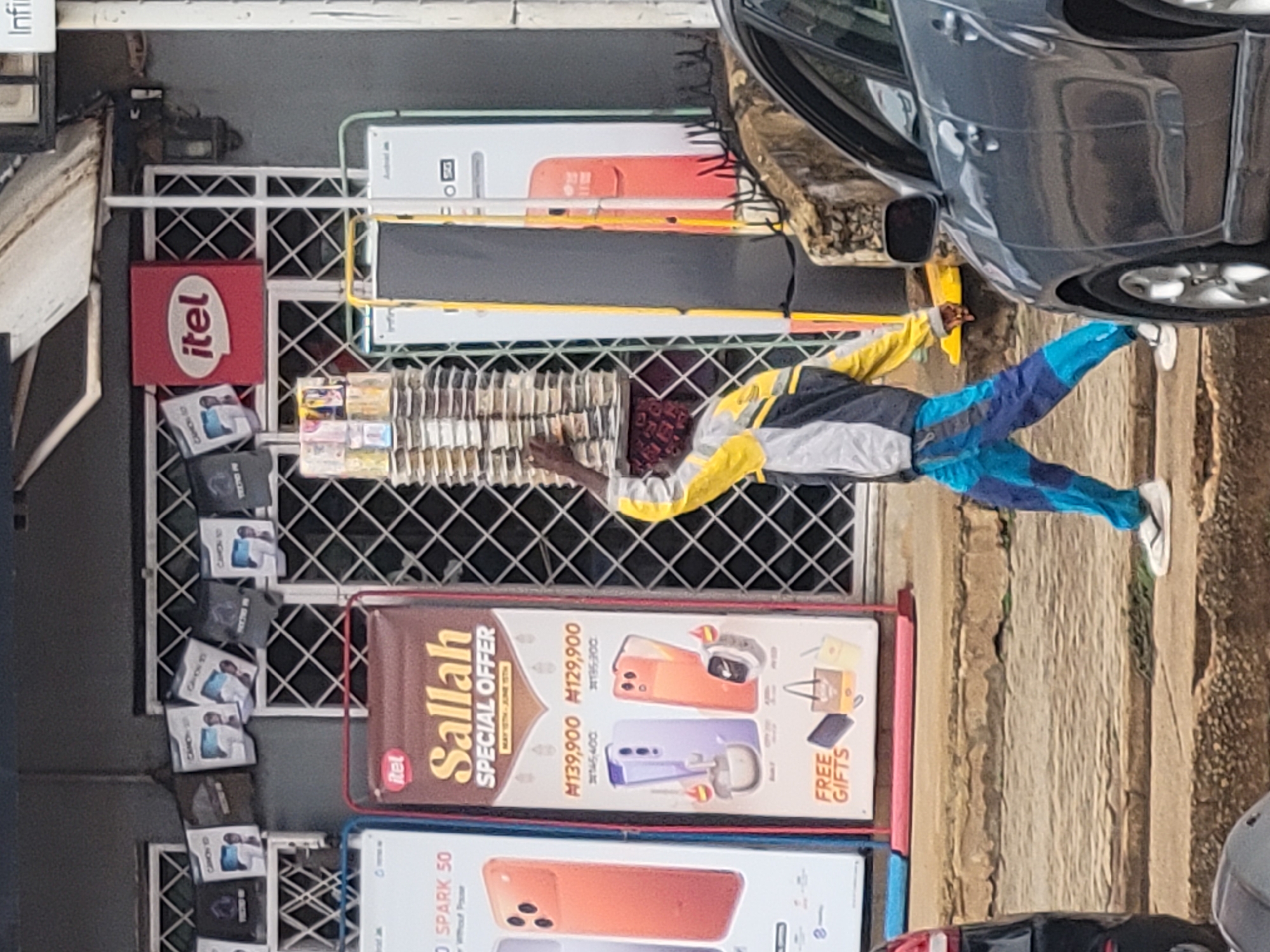 The image shows a storefront in Jos, Nigeria, featuring advertisements for Itel smartphones. The storefront displays signs for "Itel Sallah Special Offer" and the "Itel Spark 50" smartphone. There are also Canon-branded products displayed on the facade. A man is positioned near the storefront, with part of a vehicle visible on the right side of the frame. The setting appears to be an urban environment.