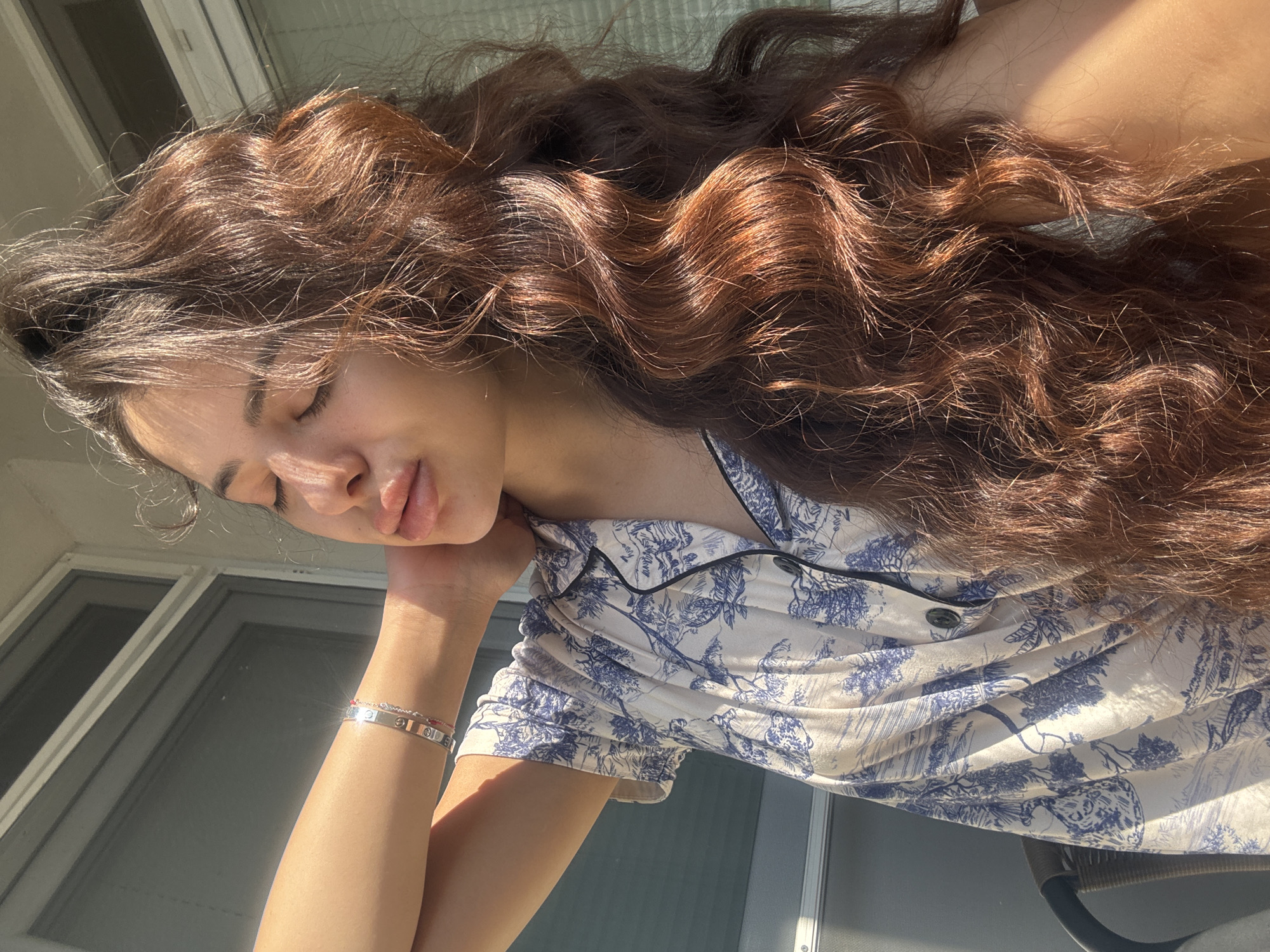 A young woman with long, wavy brown hair and closed eyes is sitting outdoors, bathed in sunlight. She is wearing a short-sleeved pajama top with a blue and white toile pattern and a silver bracelet on her left wrist. Her right arm is raised, with her hand behind her neck. The background shows a window with horizontal blinds and a neutral-colored wall. The image does not provide any specific location details within Plovdiv, Bulgaria.