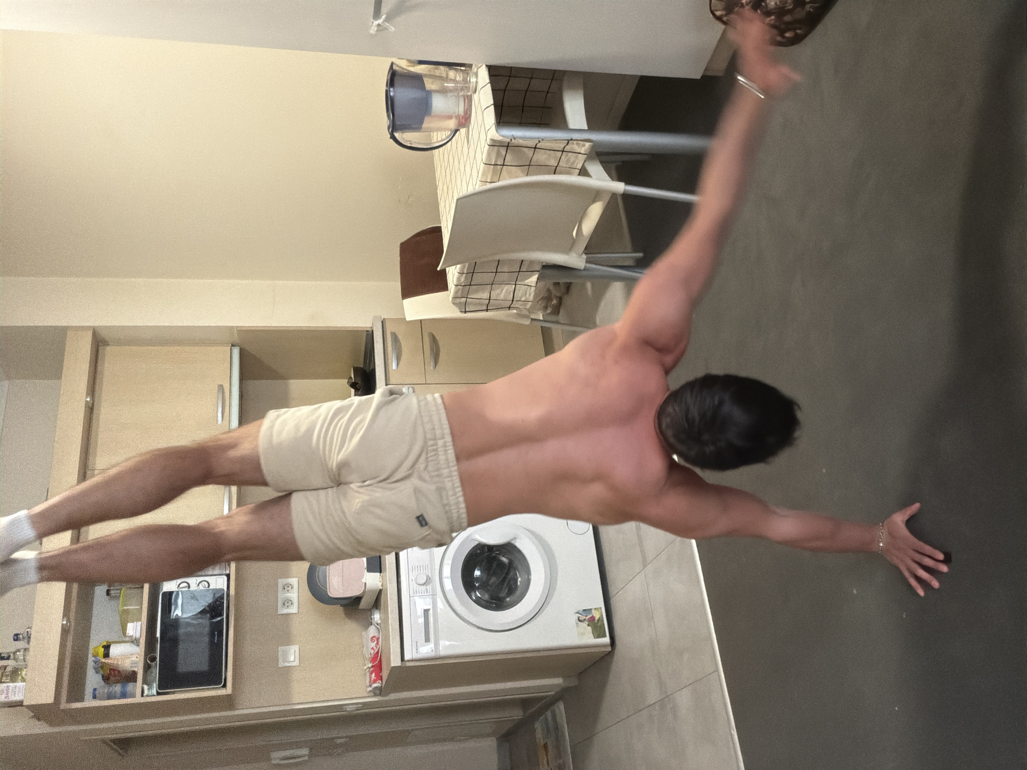 This image, presented sideways, depicts a shirtless male performing a challenging calisthenics pose, likely a human flag or a suspended plank variation. He is positioned horizontally across the frame, showcasing a muscular physique with well-defined back, arm, and leg muscles. He wears light-colored athletic shorts and white socks, with his limbs extended, suggesting significant core strength and balance.
The background is an indoor domestic setting, appearing to be a kitchen or utility area. When viewed with the correct upright orientation (rotating the image 90 degrees counter-clockwise), the person is parallel to a dark grey wall or floor, with his hands gripping the surface. To the left (which would be the main wall in an upright view) are light-colored kitchen cabinets, one of which houses a microwave oven with the partially visible brand "Homa." Below the cabinets is a white front-loading washing machine. Further in the background, a dining area is visible with a table covered by a light-colored checkered tablecloth and a couple of simple chairs. A water filter pitcher sits on the table. Other kitchen items, including bottles on shelves and an electrical outlet, are also visible.
The activity is a demonstration of advanced bodyweight strength and control. The lighting is artificial, illuminating the scene and casting some shadows. The time of day is not discernible. The location context is Sofia, Bulgaria.