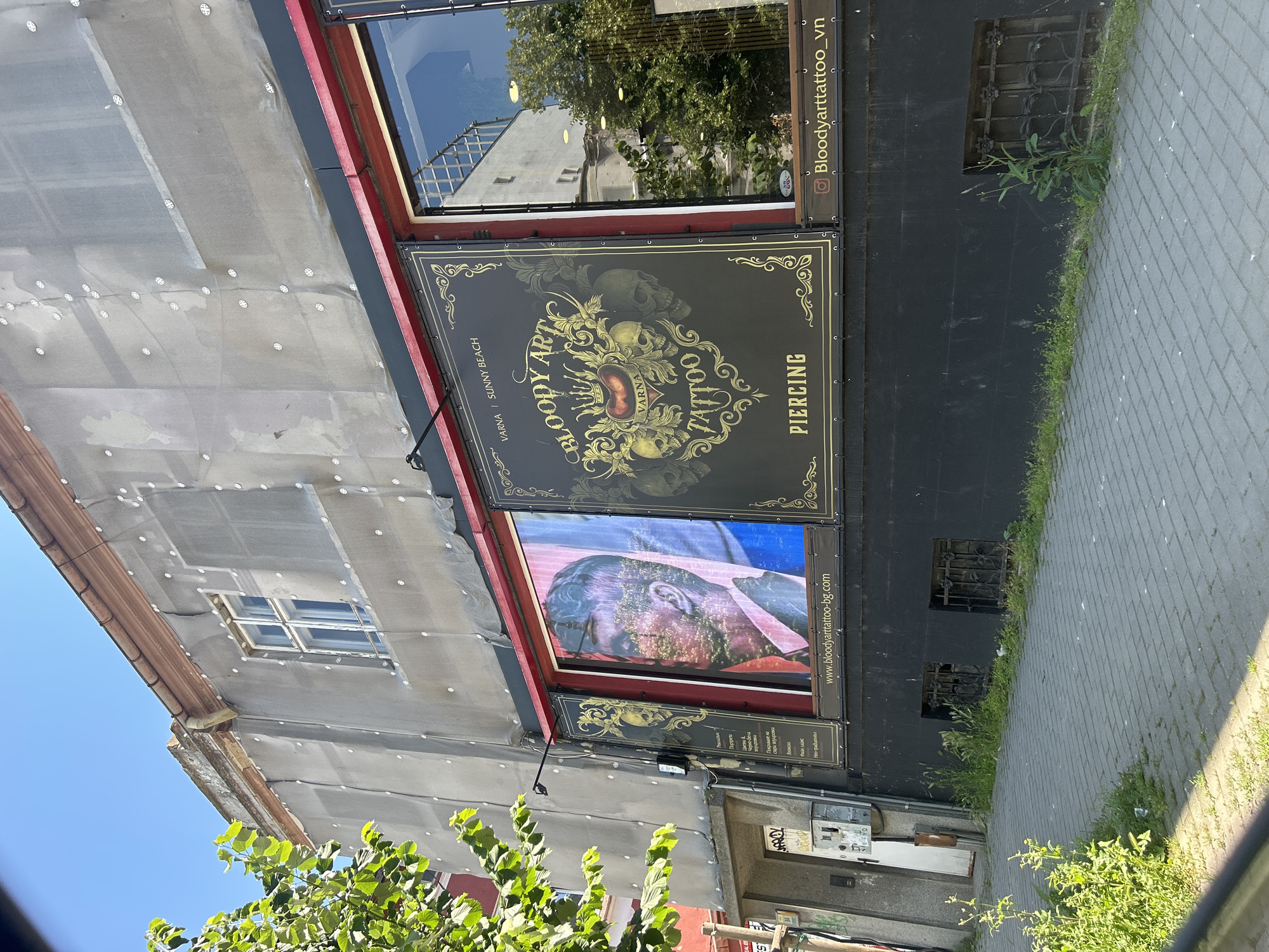 The image shows the exterior of a tattoo and piercing shop located in Varna, Bulgaria. The shop, named "Bloody Art Tattoo," is situated on the ground floor of a building covered in gray sheeting and adorned with white circular fasteners, suggesting it may be undergoing renovation or maintenance. Large display posters are visible on the storefront. One poster features a stylized design with skulls, a crown, and the text "BLOODY ART TATTOO VARNA SUNNY BEACH" and "PIERCING." Another poster displays a close-up portrait of a man with a double exposure effect. Below the posters, the shop has a black facade and barred windows. A sidewalk with a cobblestone pattern runs in front of the establishment, with overgrown grass along the building's base. The sky is clear and blue, indicating a sunny day.