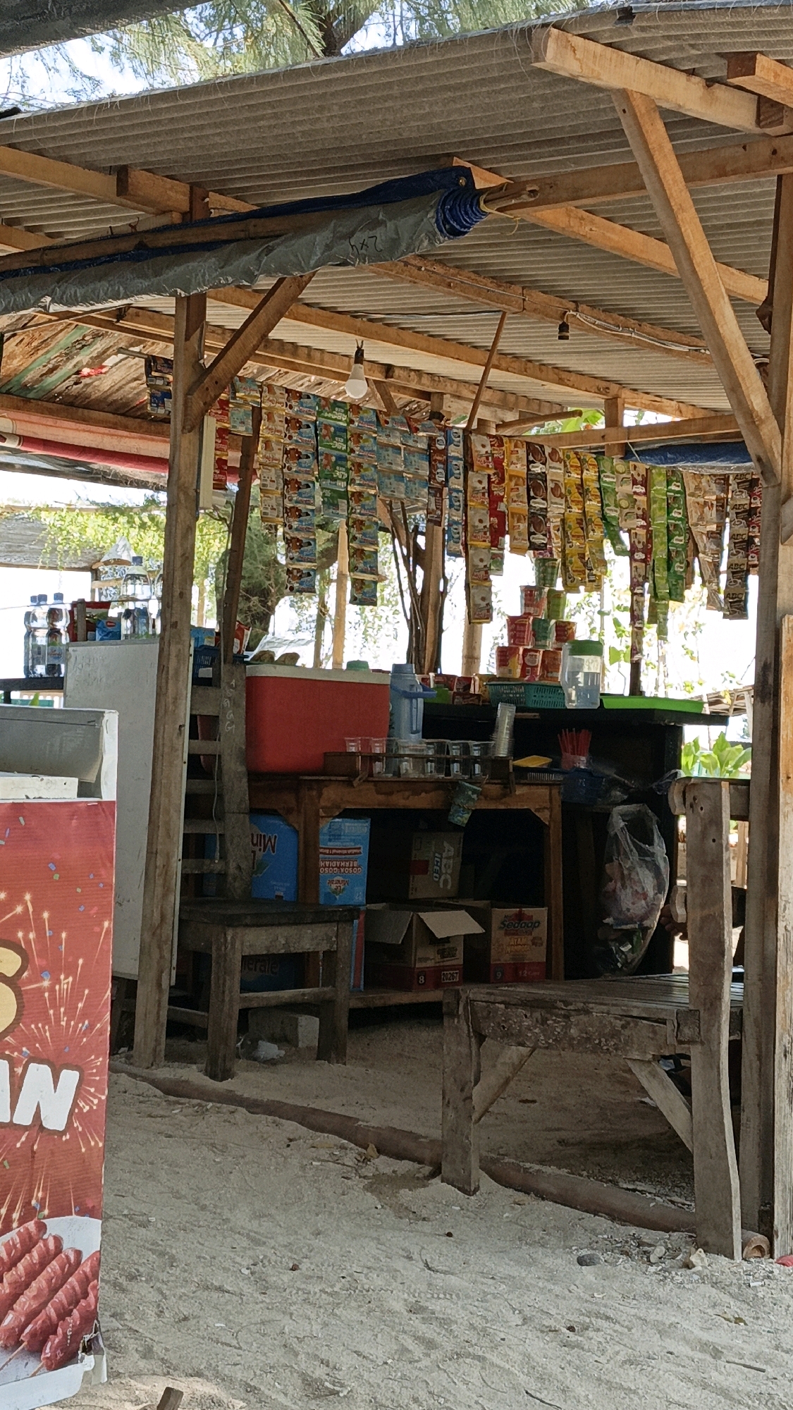 The image depicts an outdoor stall, likely a small convenience store or snack stand, in Cikal, Indonesia. The stall is constructed from rough wooden beams and covered with a corrugated metal roof. Numerous small packets of snacks or instant food are hanging in rows from the ceiling, displaying a variety of colorful packaging.
Below the hanging goods, a wooden counter displays several items, including a red cooler, bottles of water, stacks of small cups, and what appear to be packets of seasoning or instant coffee. There are also several cardboard boxes, some open, on shelves beneath the counter. A blue plastic crate is visible on the ground, along with a shopping bag containing plastic items.
To the left of the stall, a large red and white advertisement banner featuring an image of sausages on skewers is partially visible. The ground is covered with sand and small pebbles. In the background, the light suggests a bright, sunny day, with trees and foliage visible beyond the stall. The overall impression is that of a casual, open-air market or roadside vendor.